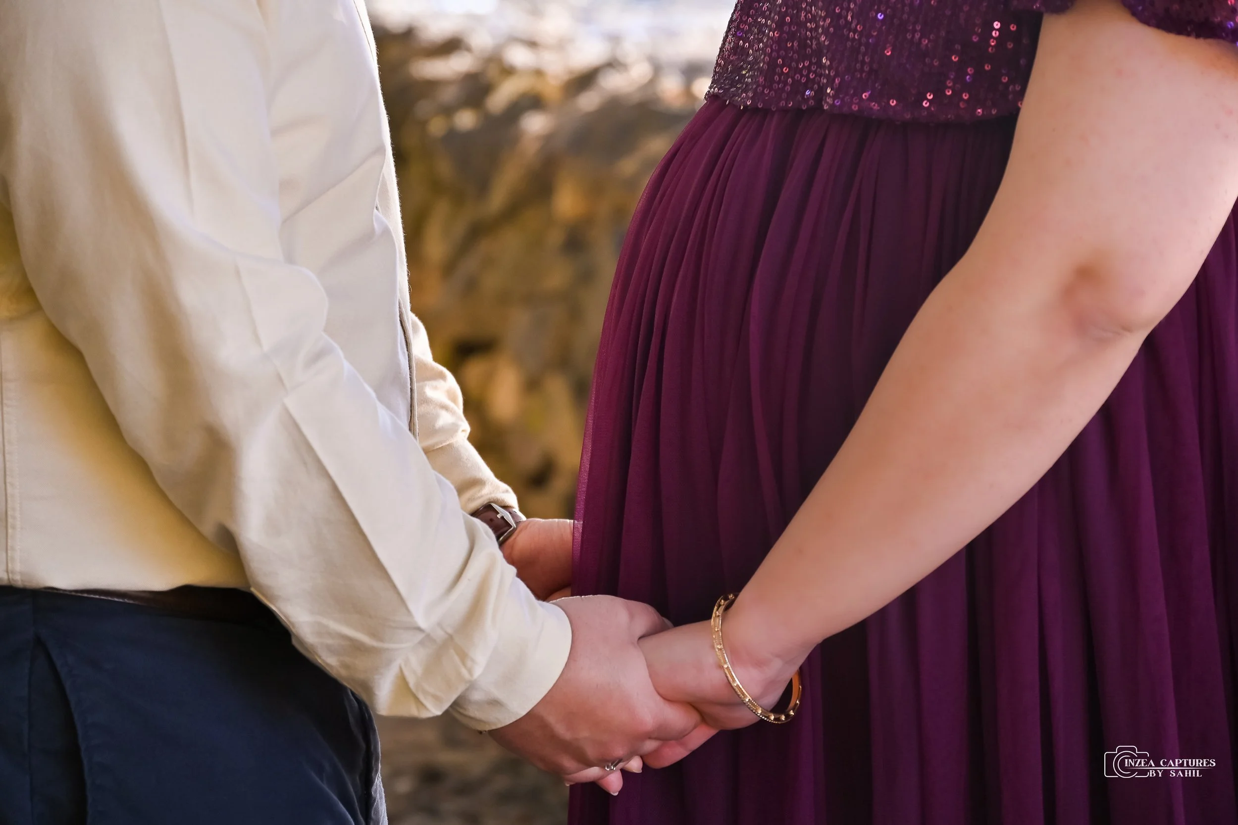 A couple holding hands, with the woman wearing a purple dress and the man wearing a beige shirt. The focus is on their hands and the woman's arm with a bracelet.