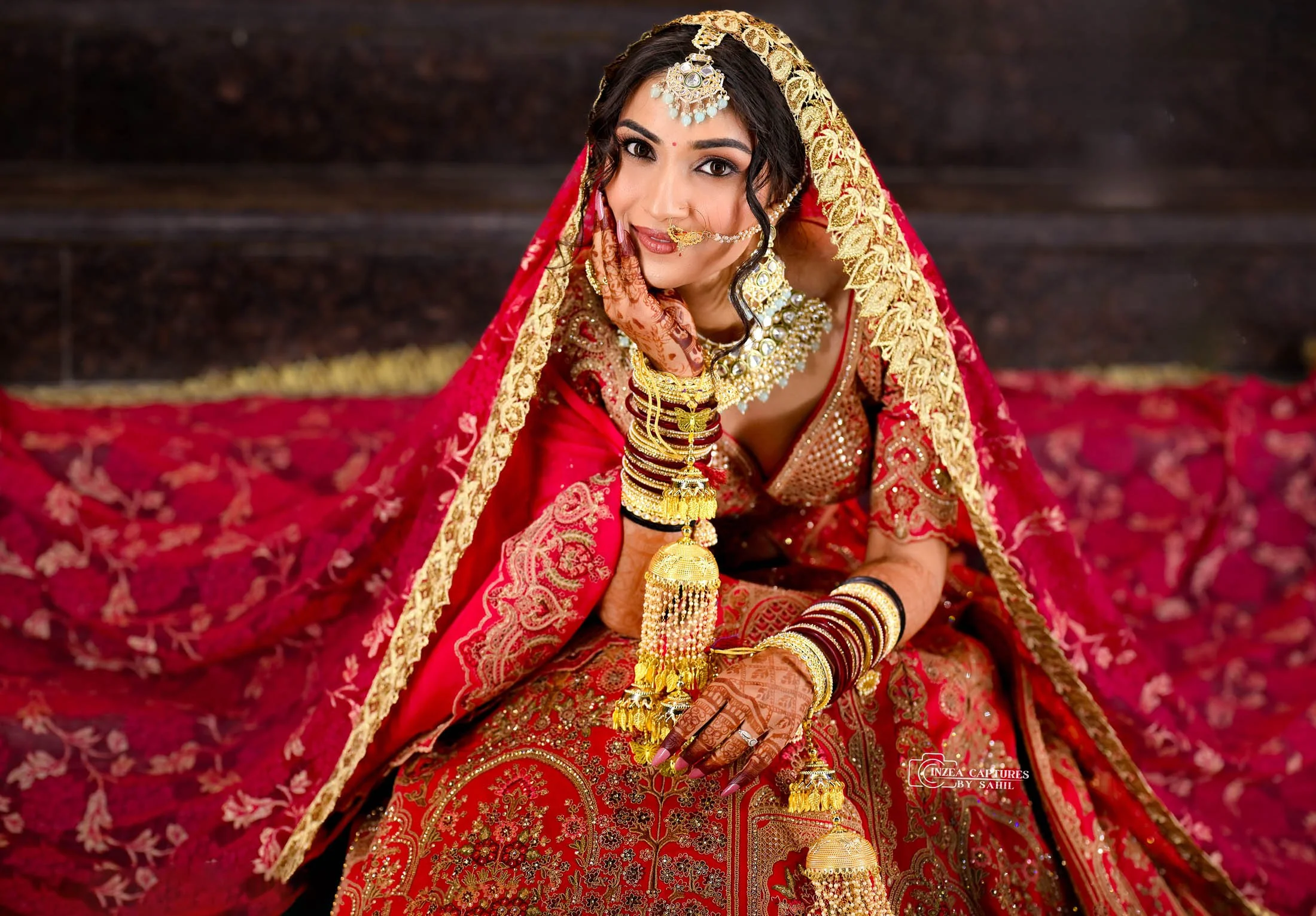 An Indian woman dressed in traditional red and gold bridal attire, wearing ornate jewelry including necklaces, bangles, earrings, and a nose ring, sitting on a red embroidered carpet with a wooden background.
