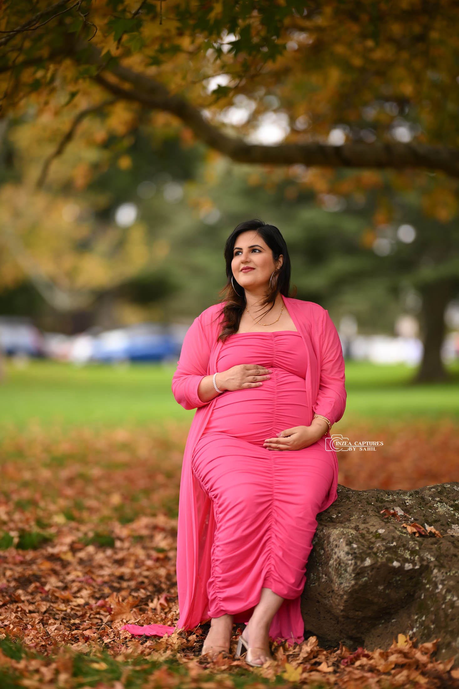 A pregnant woman dressed in a pink outfit, sitting on a rock in a park with autumn leaves on the ground, surrounded by trees with fall foliage.
