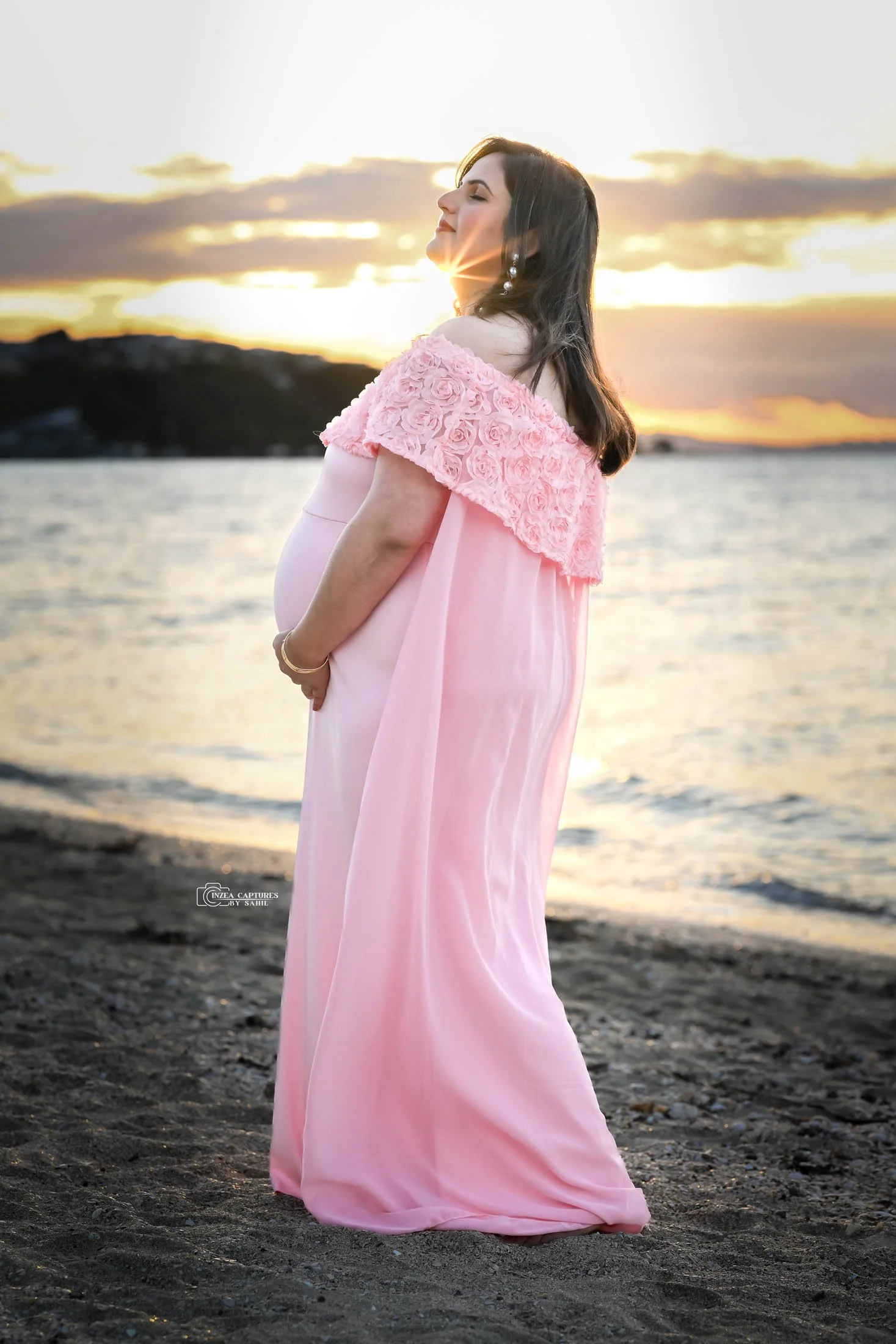 A pregnant woman in a pink dress with floral embellishments on the shoulder stands on a beach during sunset, gently holding her belly with her eyes closed and a serene expression.
