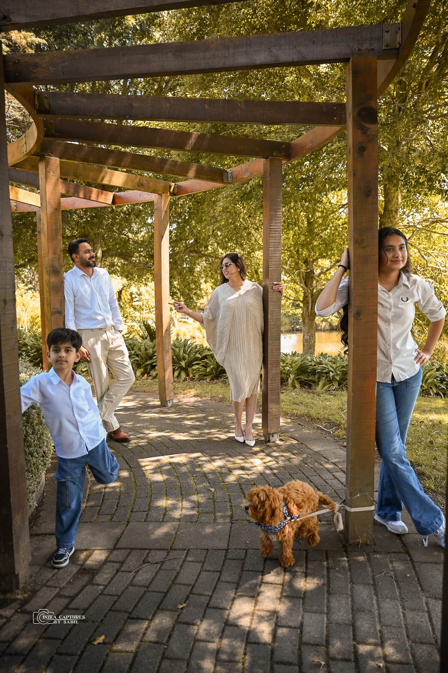 A family of five, including a man, woman, teenage girl, young boy, and a small dog, standing and posing outdoors under a wooden structure in a park with trees and a body of water in the background.