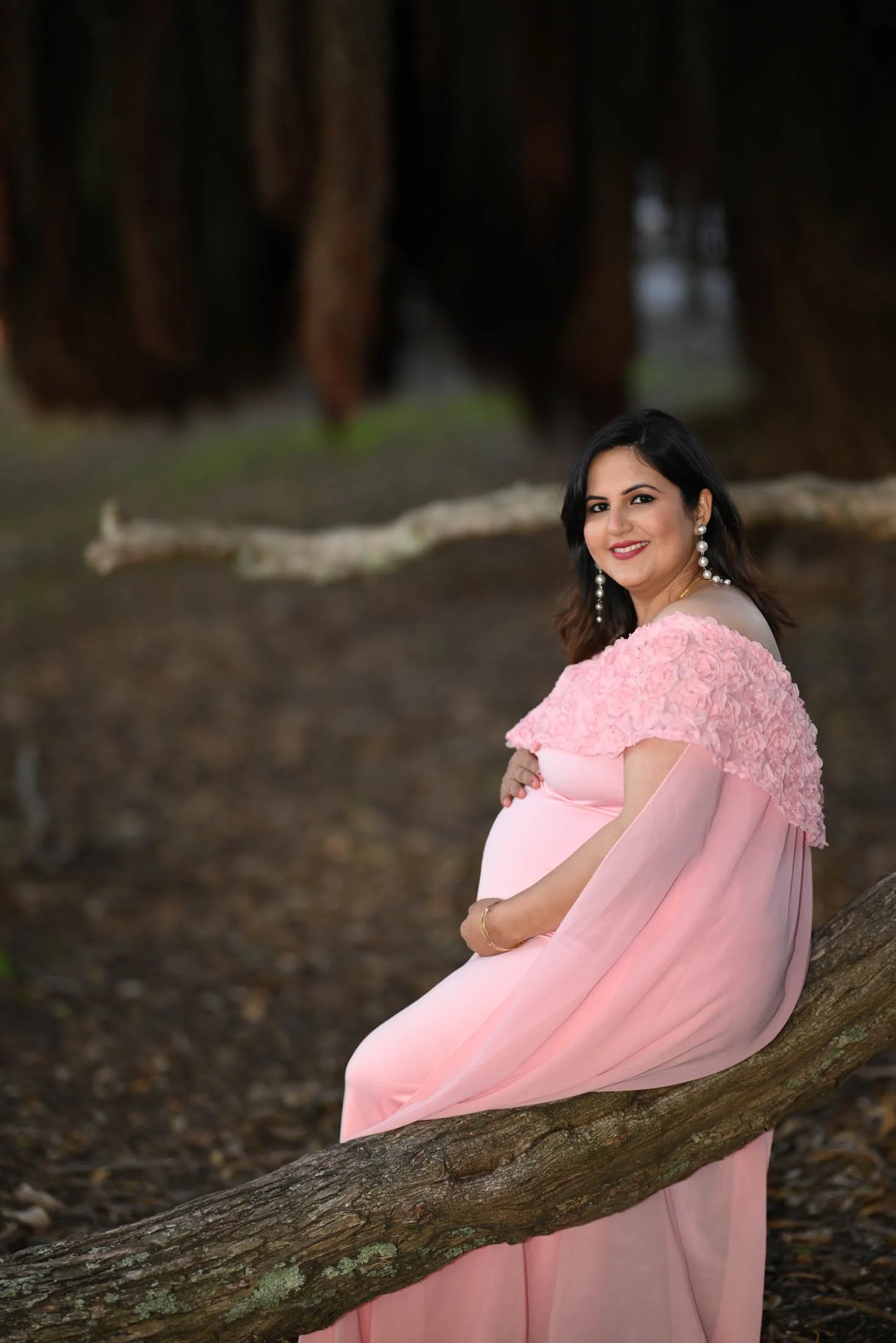 A pregnant woman in a pink dress sitting on a tree branch in a forest, smiling at the camera, wearing earrings and a bracelet.