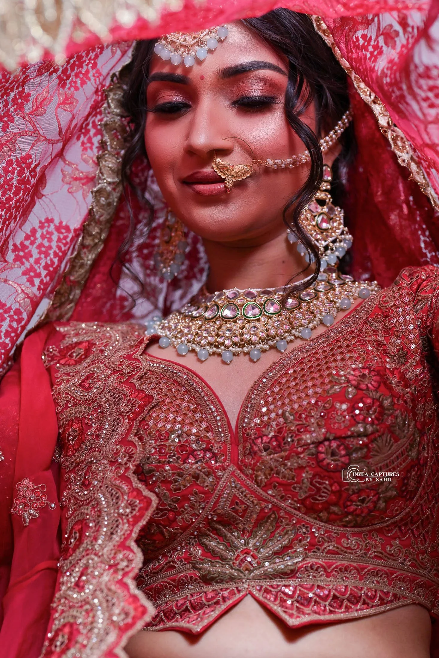 A woman dressed in traditional Indian bridal attire wearing a red embroidered lehenga and ornate jewelry including a necklace, earrings, and a nose ring with chain. She has a red veil partially covering her head and closed eyes.