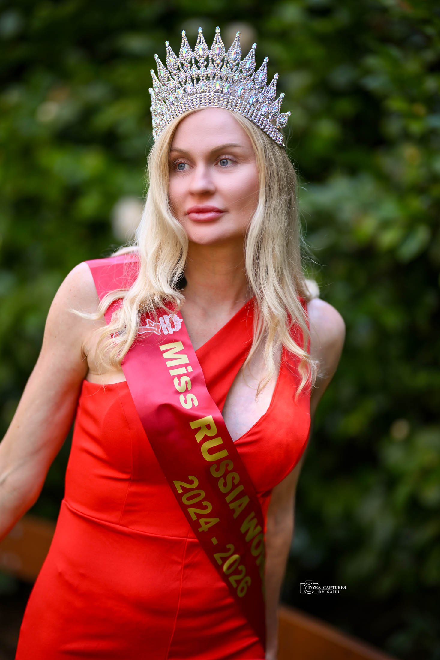 A woman with long blonde hair wearing a crown and a red dress with a sash reading "Miss Russia 2024-2025" standing outdoors with green foliage in the background.