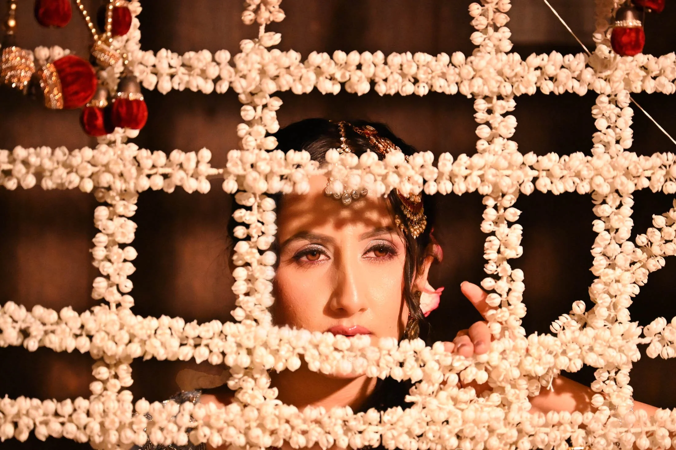 A woman with traditional jewelry and makeup looking through a decorative lattice made of white flowers, with Christmas ornaments hanging around.