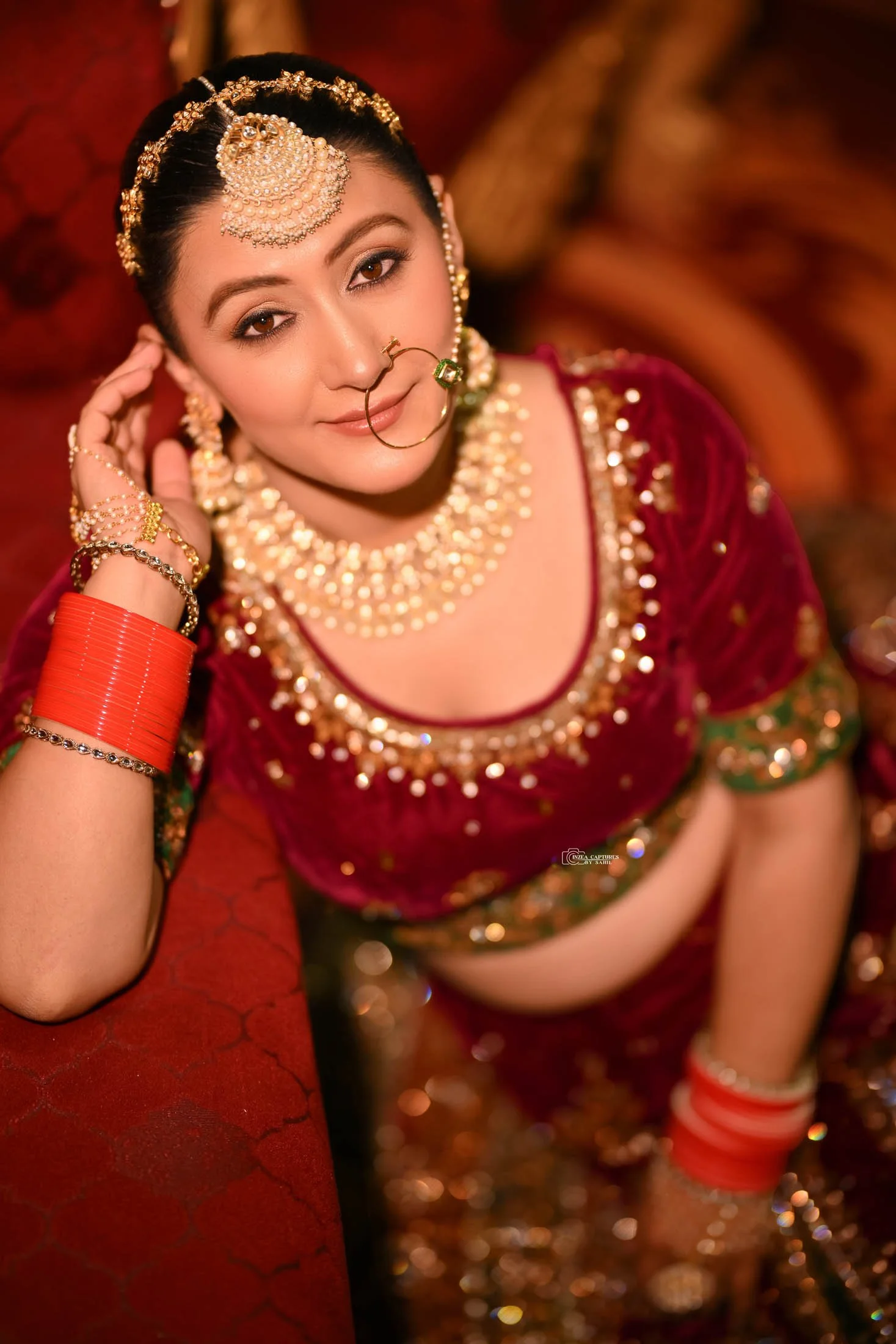 A woman dressed in traditional Indian bridal attire with elaborate jewelry, including a large maang tikka, earrings, nose ring, necklace, and bangles, smiling gently and resting her head on her hand.