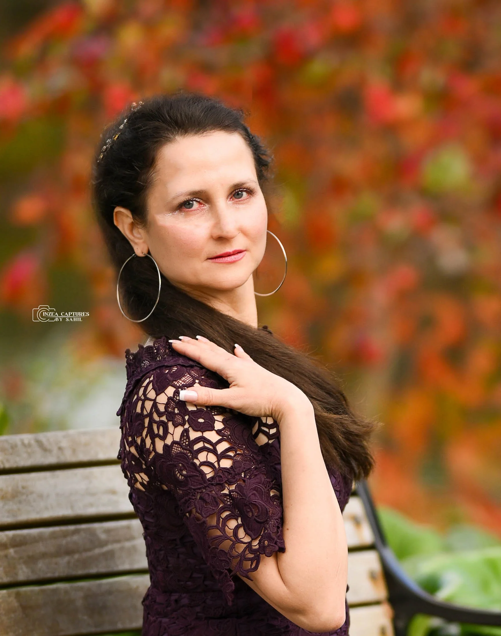 A woman with dark hair, wearing large hoop earrings and a purple lace dress, sitting on a park bench with a background of fall-colored trees.