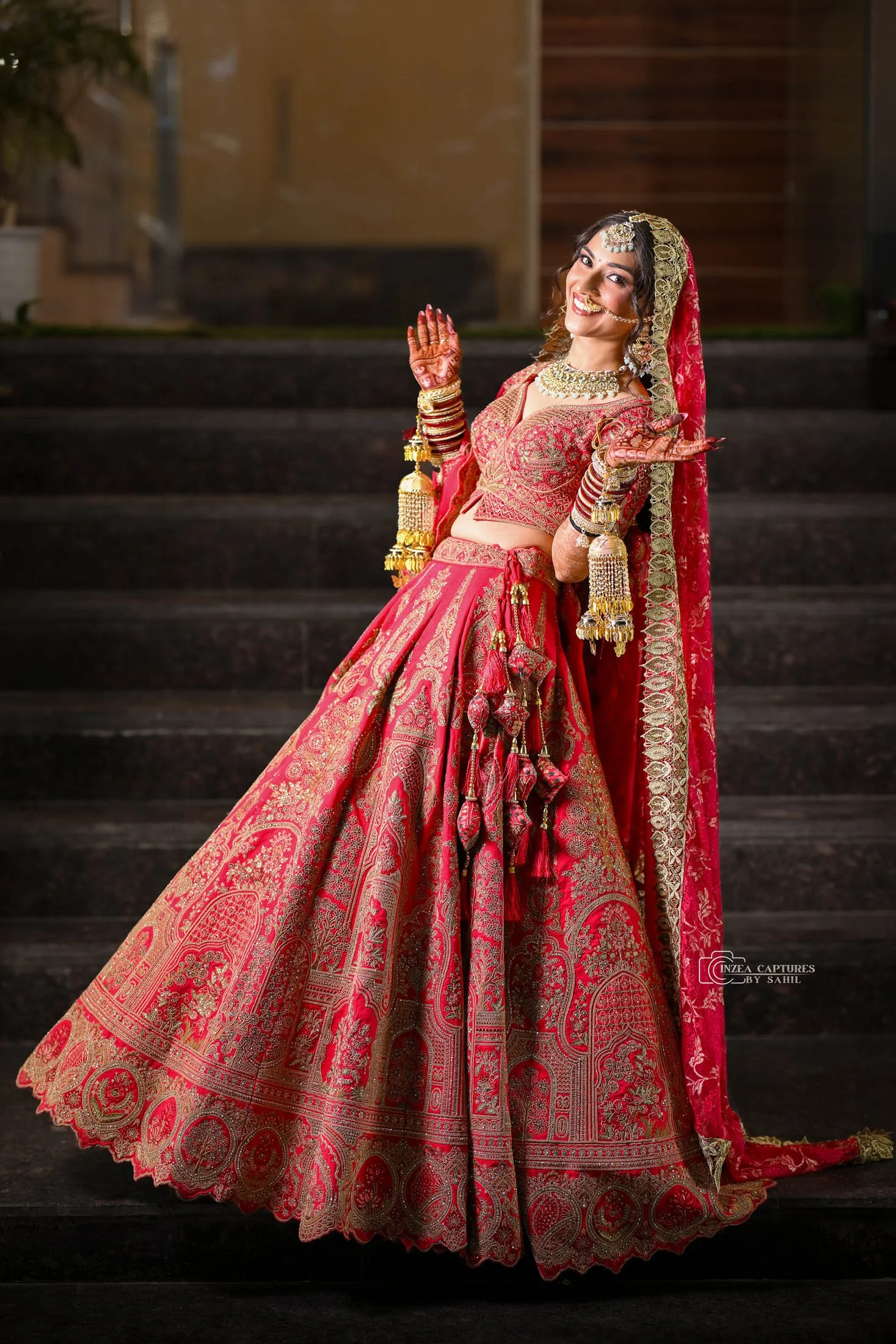 A woman in a traditional red and gold bridal outfit, adorned with jewelry and bangles, smiling and posing on stairs during a wedding ceremony.