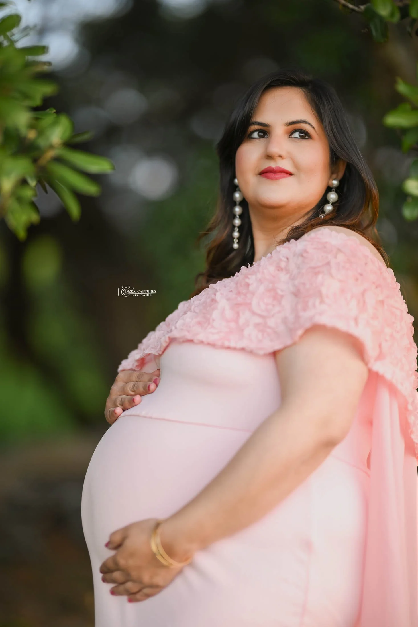 A pregnant woman wearing a pink dress with lace embellishments, standing outdoors with greenery in the background. She is looking to the side, with her right hand protecting her belly and her left hand resting below her belly. She has dark, shoulder-