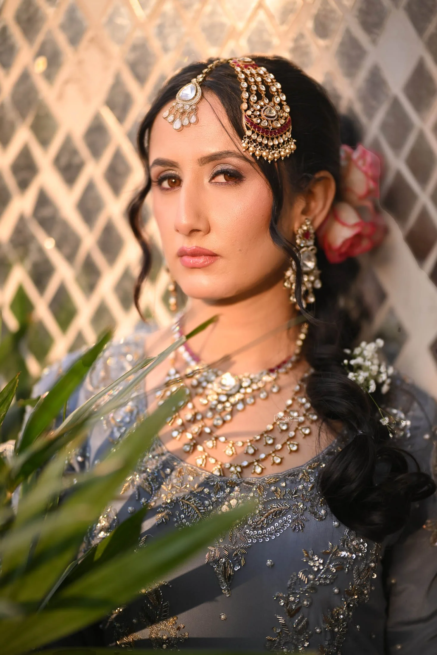 A woman dressed in traditional Indian attire, wearing elaborate jewelry and earrings, with flowers in her hair, standing in front of a lattice background, partially obscured by green leaves.