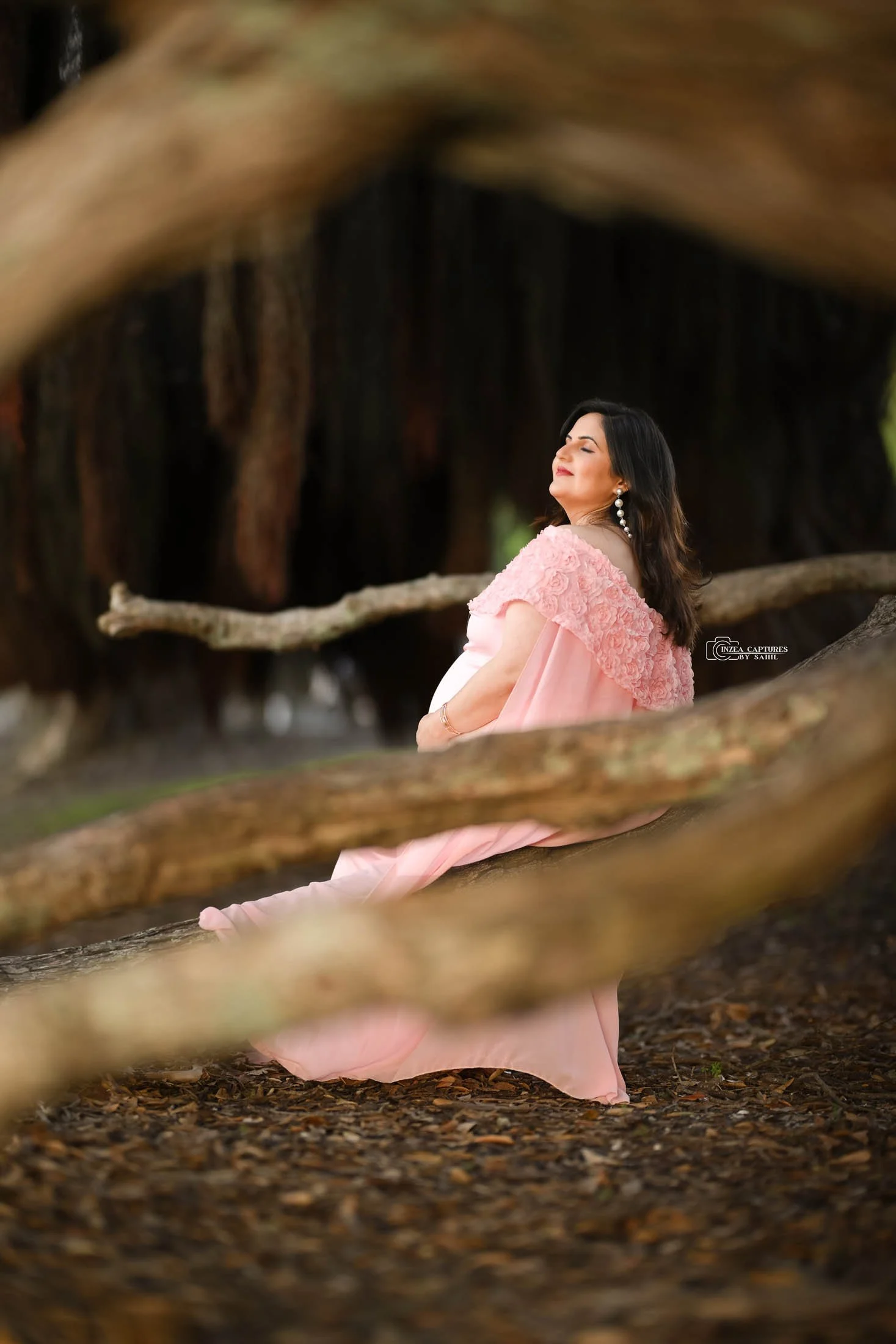 A woman in a pink dress sitting on the ground, framed by tree branches in a natural setting, with her eyes closed and a peaceful expression.