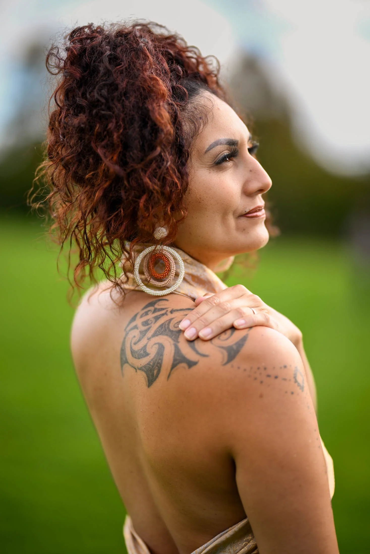 Portrait of a woman with curly red hair, large earrings, and a tattoo on her shoulder, standing outdoors with a blurred green background.