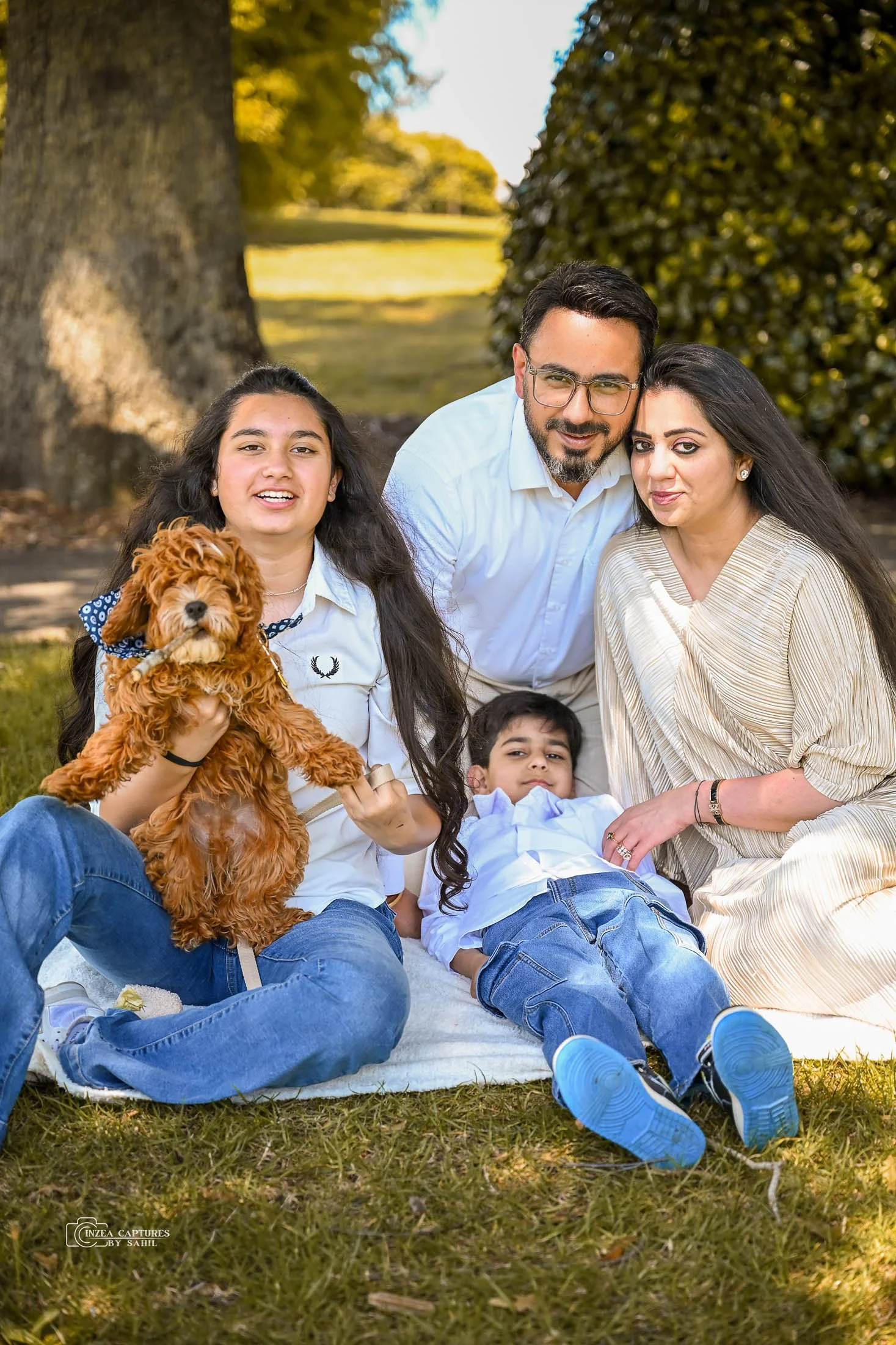 Family of five, including two adults, two children, and a dog, sitting outdoors on a blanket in a park on a sunny day.