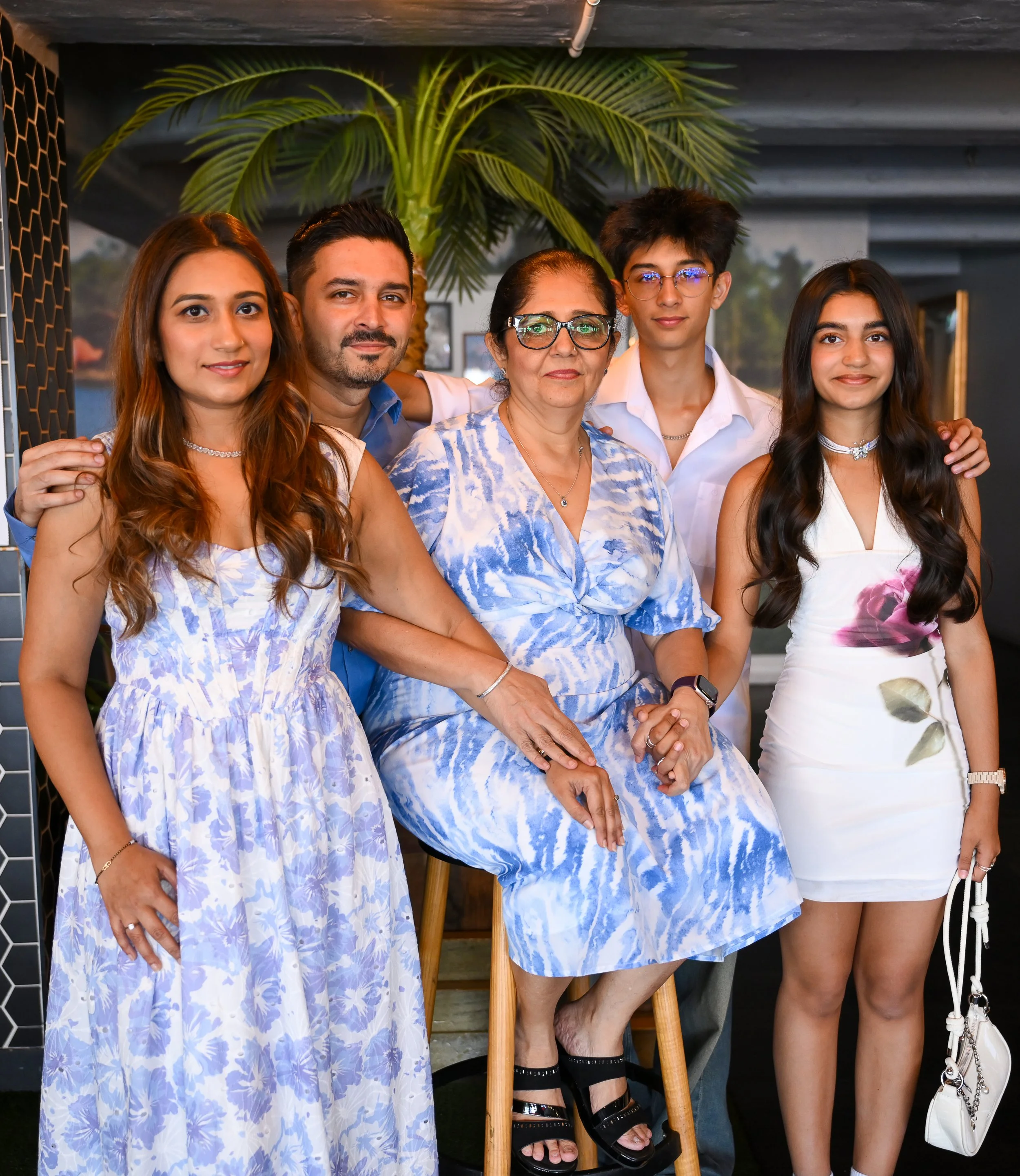 A family portrait of six people, five adults and one teenager, posing indoors in front of a large green plant, all dressed in blue and white clothing.