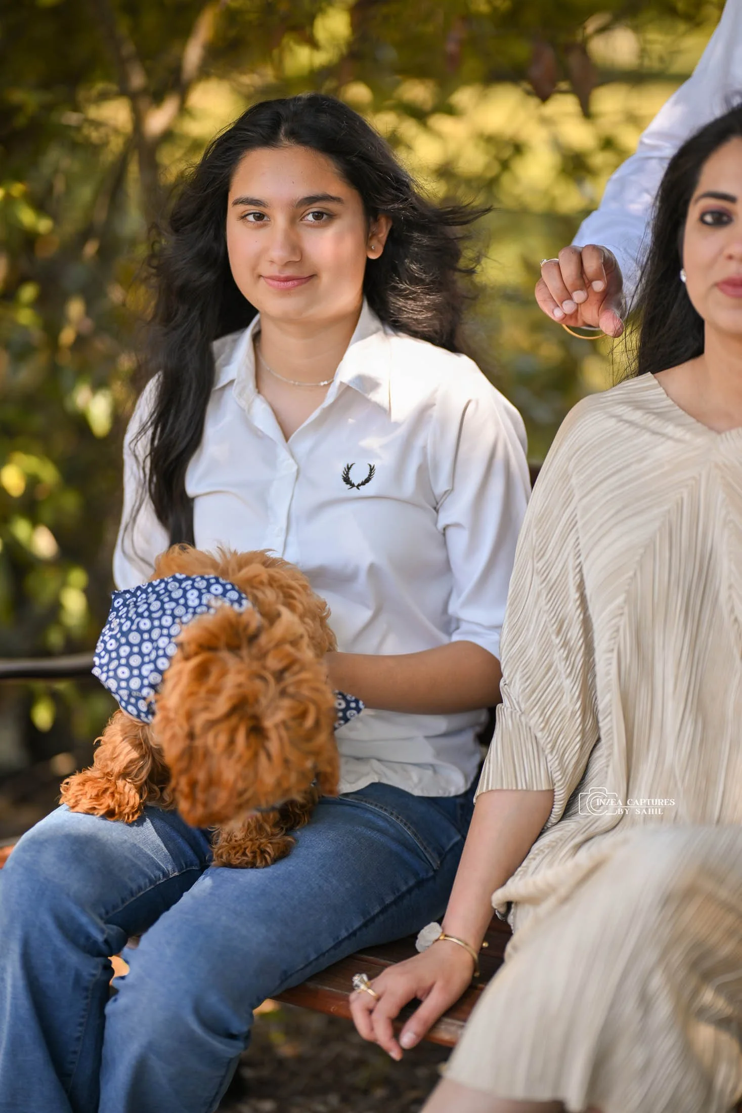Young woman with long dark hair sitting outdoors holding a small brown dog with a blue bandana, with another woman partially visible beside her.