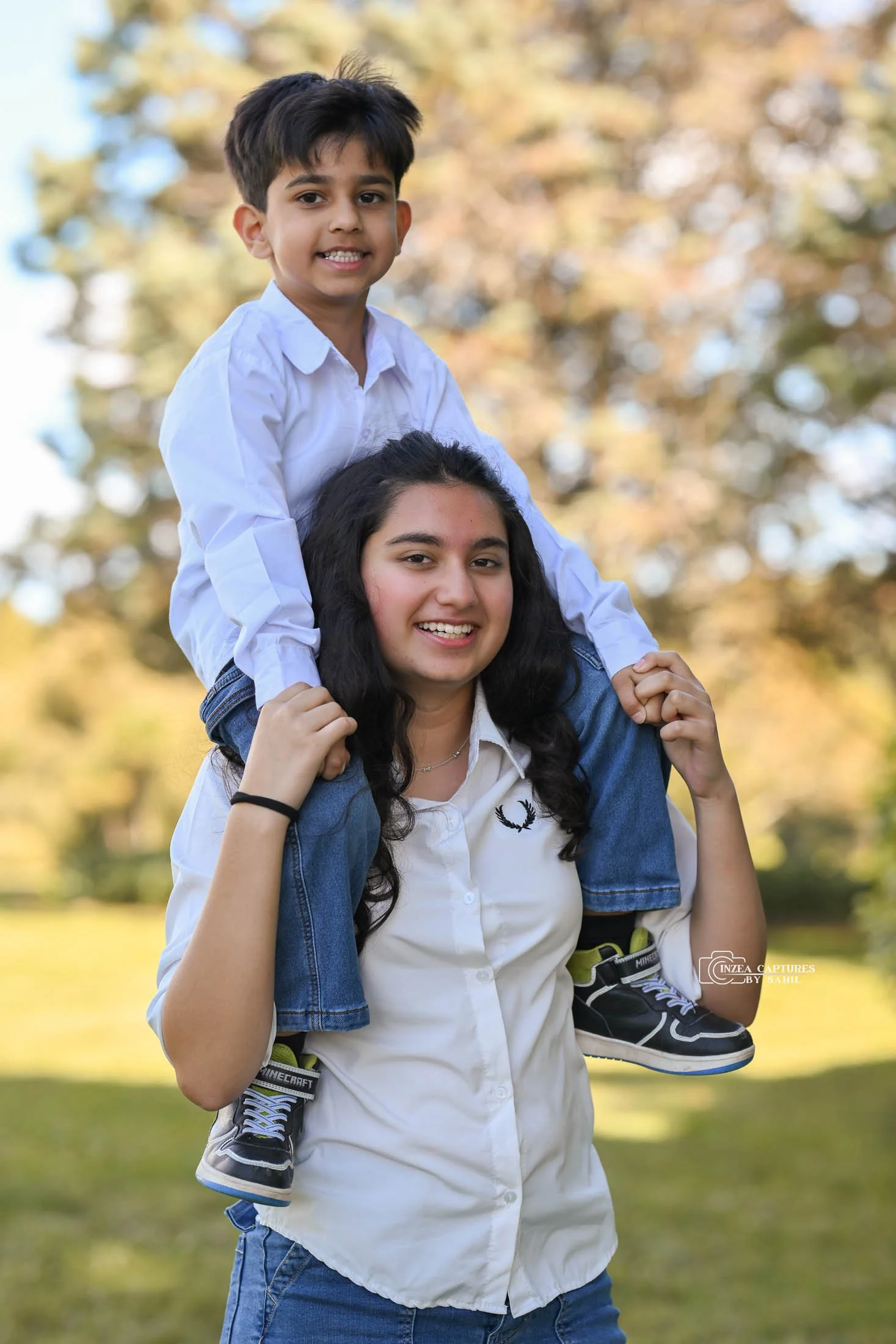 A young boy is sitting on a woman's shoulders, both smiling outdoors with trees and sunlight in the background.