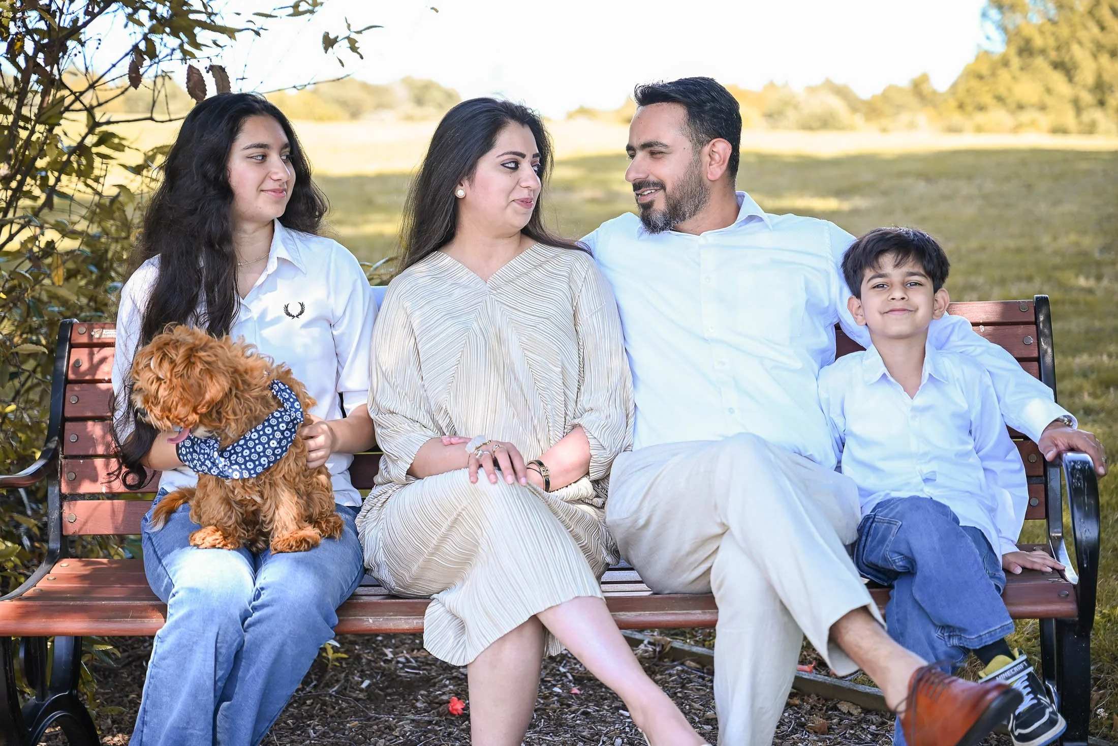 Family of four with a dog sitting on a park bench outdoors on a sunny day