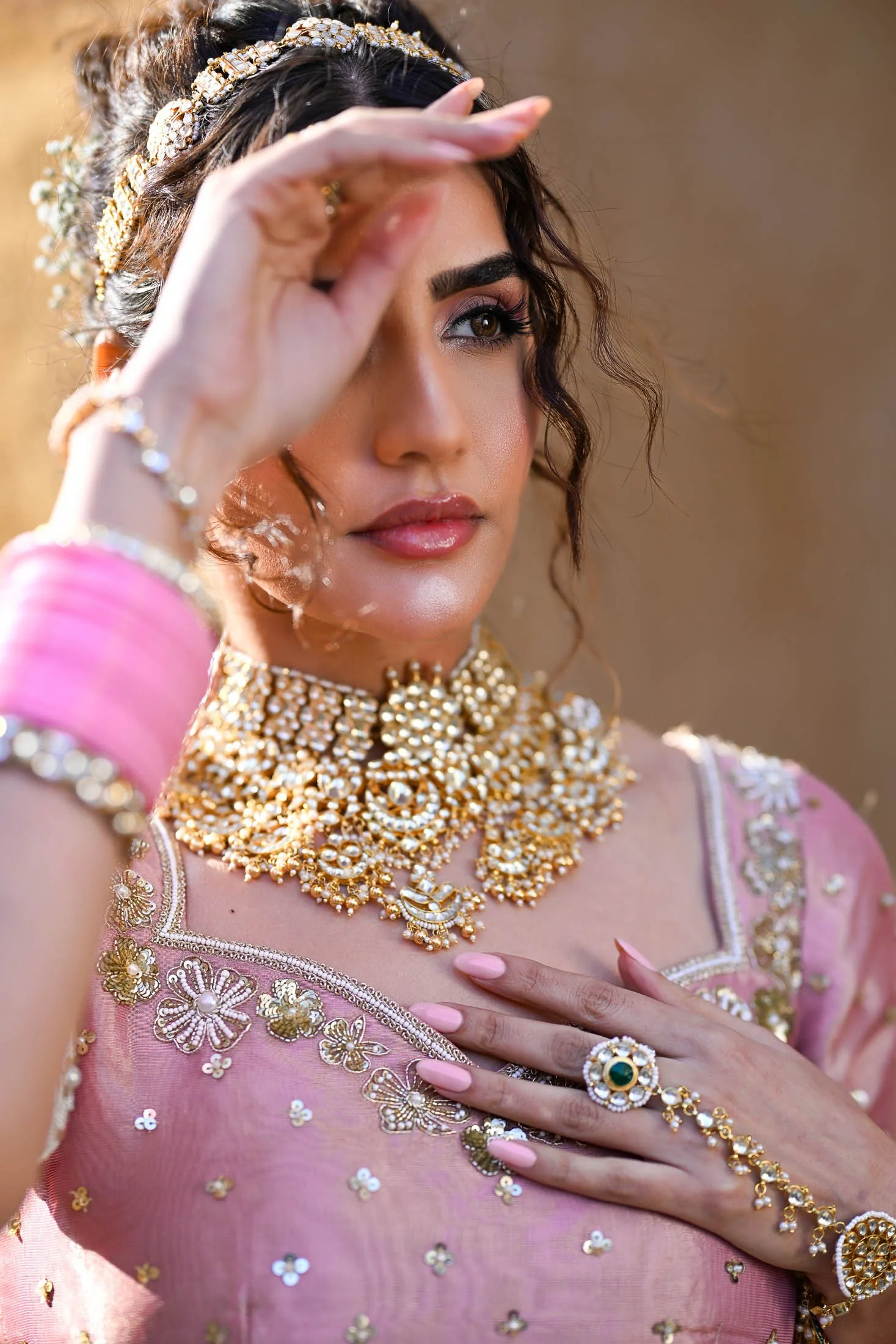 A woman dressed in traditional Indian jewelry and attire, wearing a pink saree with embroidery, with her hand touching her chest and the other near her forehead, adorned with rings, bangles, necklaces, and a headpiece.