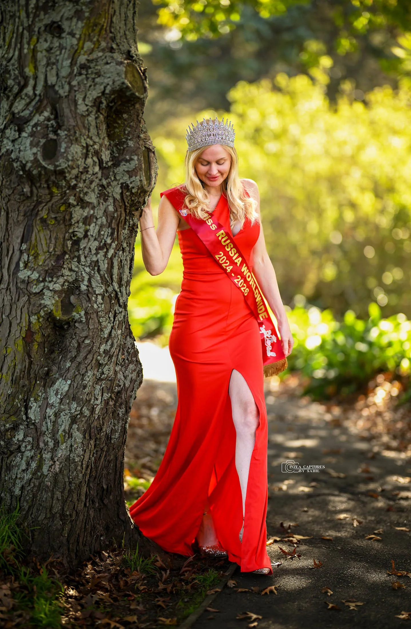 A woman dressed in a red evening gown, wearing a crown and a sash that reads 'Miss Russia Worldwide 2014-2026,' stands outdoors next to a tree with a serene expression, surrounded by a lush green background.