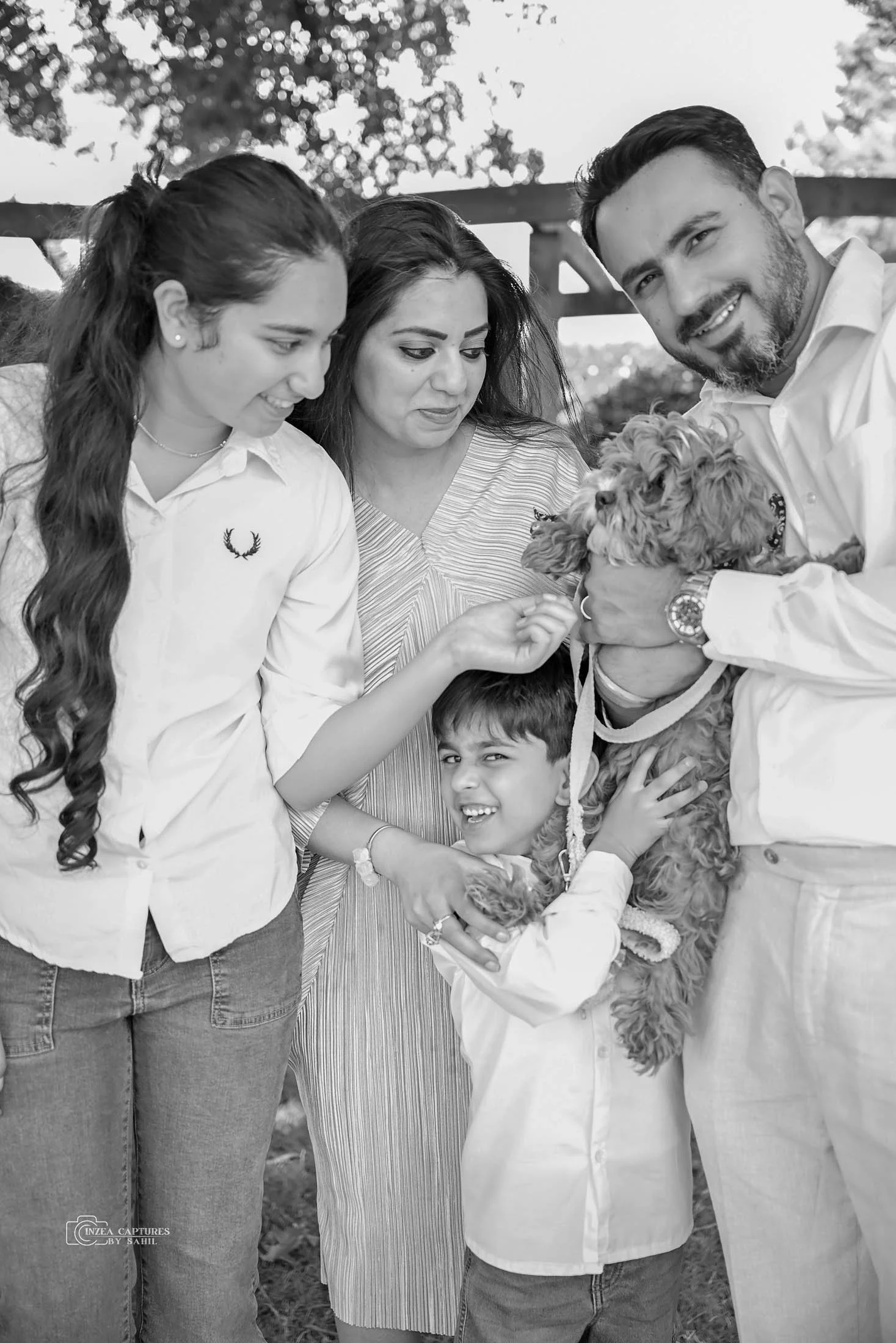 A family of five, including two adult men, two young girls, and a young boy, gathered outdoors with a small dog. All are smiling and touching the dog, celebrating a happy moment.