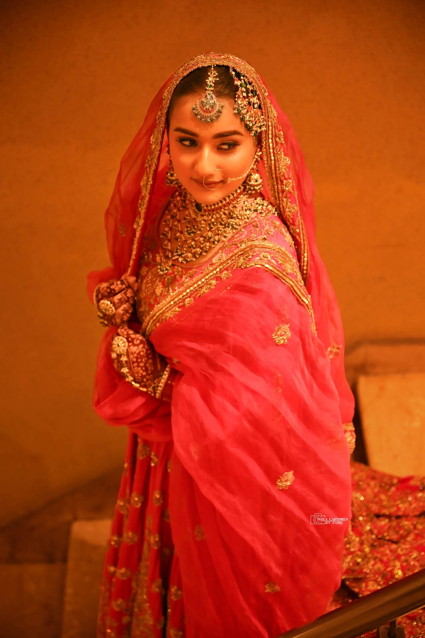 A woman in traditional Indian bridal attire with intricate jewelry and a red saree, tilting her head slightly with a soft smile.