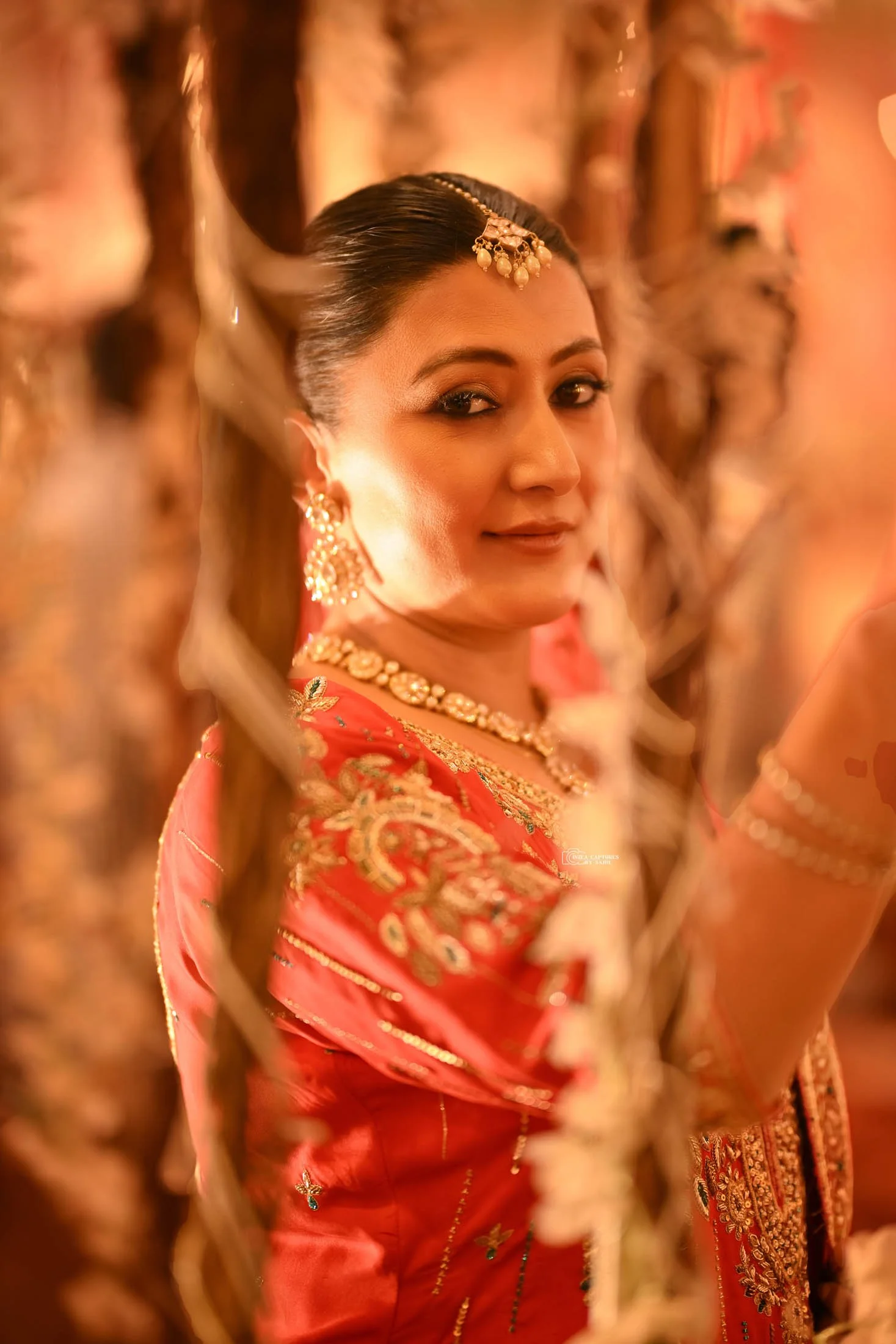 A woman dressed in traditional Indian attire, adorned with gold jewelry, smiling softly, and facing the camera through hanging floral decorations.