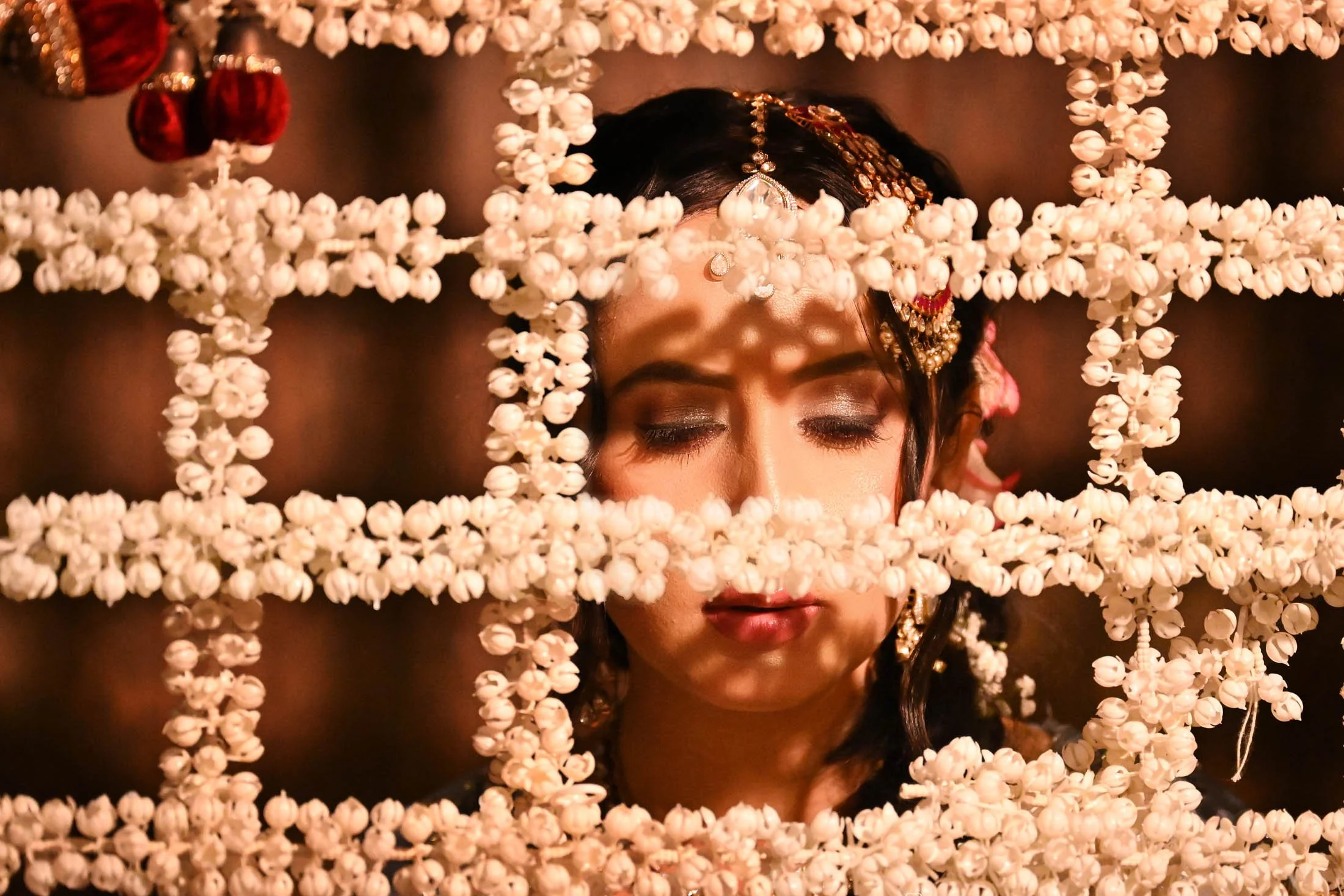 A woman with traditional Indian jewelry and makeup, with her eyes closed, framed by strings of white flowers.