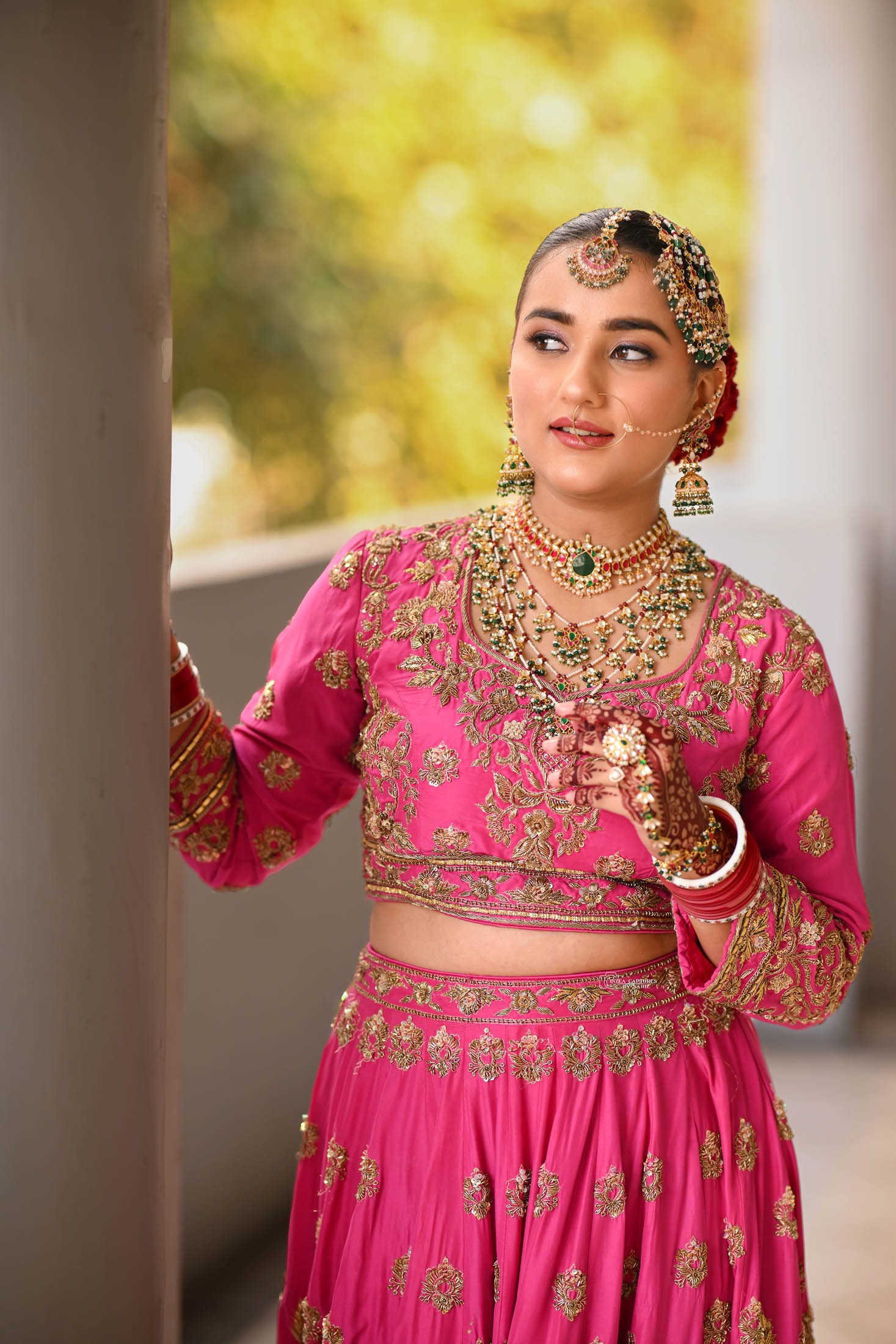 A woman dressed in traditional Indian bridal attire, wearing a vibrant pink embroidered lehenga with gold details, and adorned with multiple necklaces, earrings, bangles, and a nose ring. She has henna on her hand and is standing outdoors, looking to
