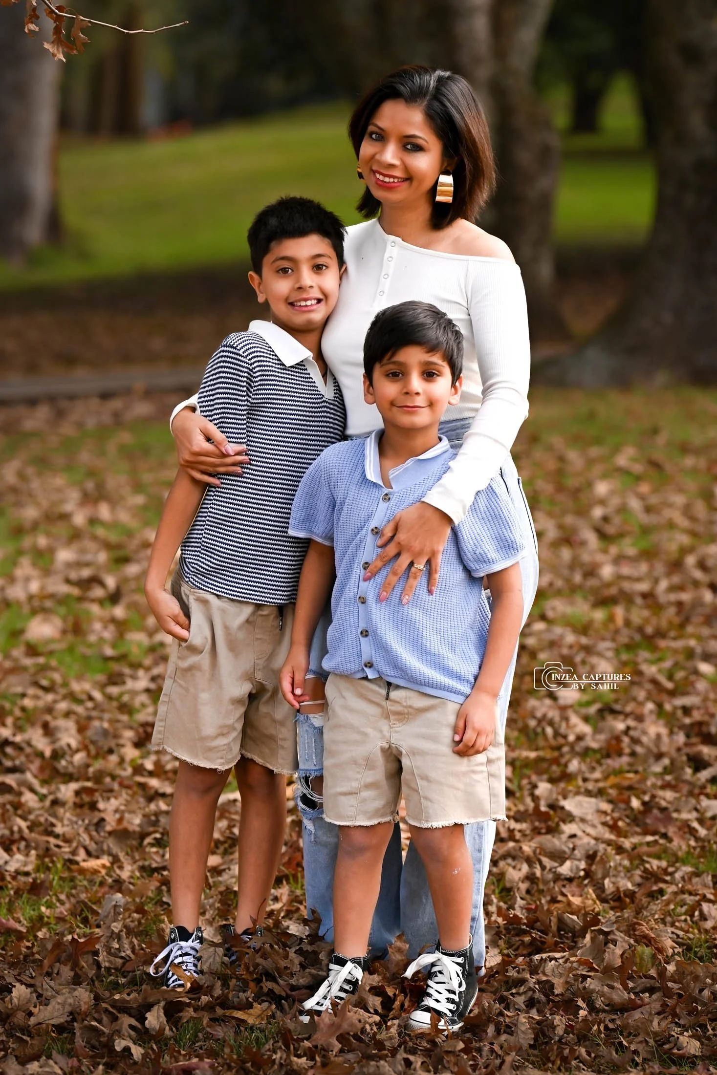A woman and two young boys standing outdoors on fallen leaves in a park during autumn. The woman has shoulder-length dark hair and is wearing a white long-sleeve top and large hoop earrings. The boys are wearing casual shirts and khaki shorts, with s