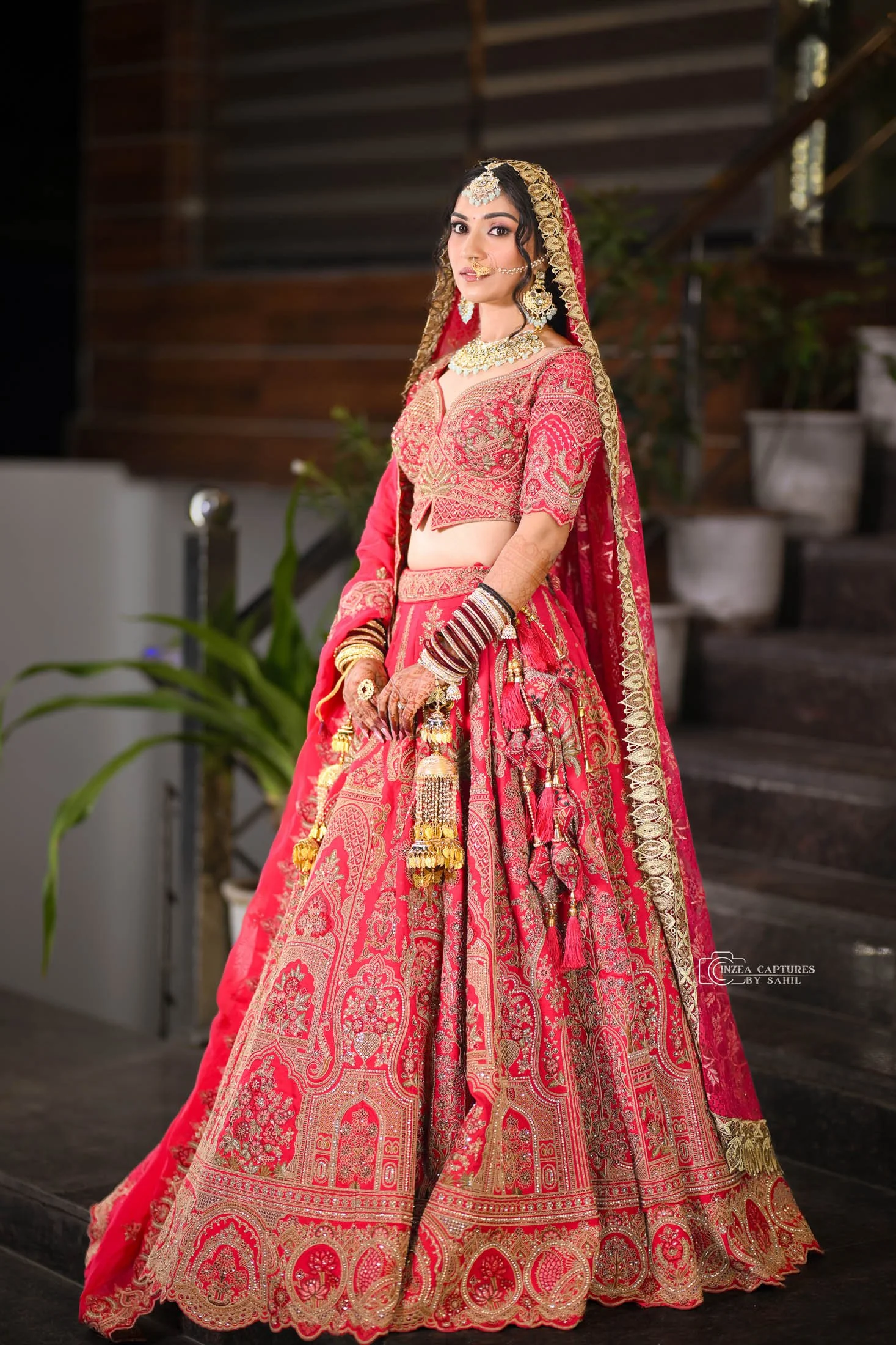 A woman dressed in traditional red bridal attire with gold embroidery, wearing jewelry including a headpiece, necklace, earrings, nose ring, bangles, and henna on her hands, standing indoors with wooden stairs and potted plants in the background.