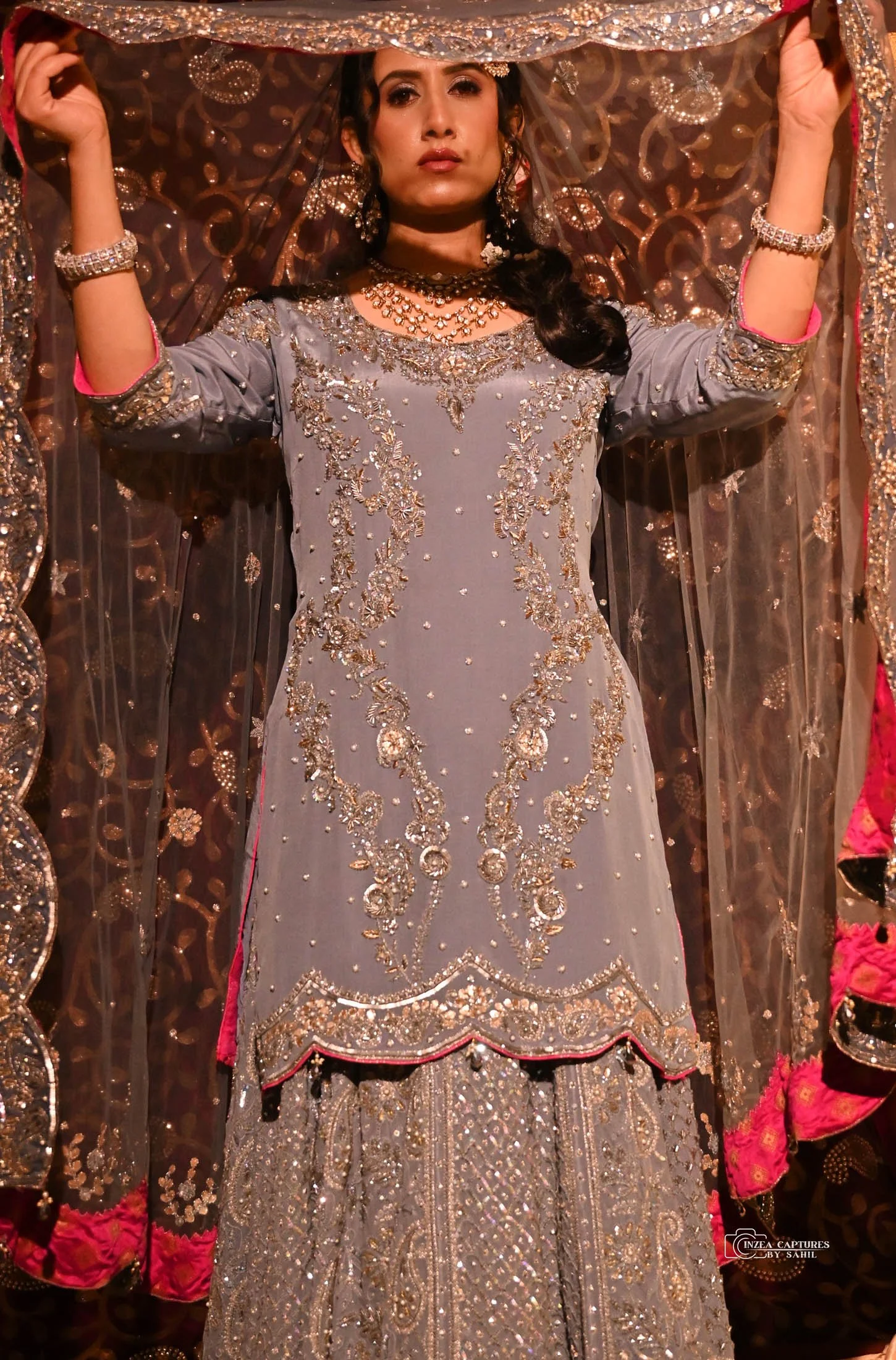 A woman dressed in ornate, traditional South Asian wedding attire, including a heavily embroidered dress with silver and gold embellishments, standing in front of a matching decorated backdrop.