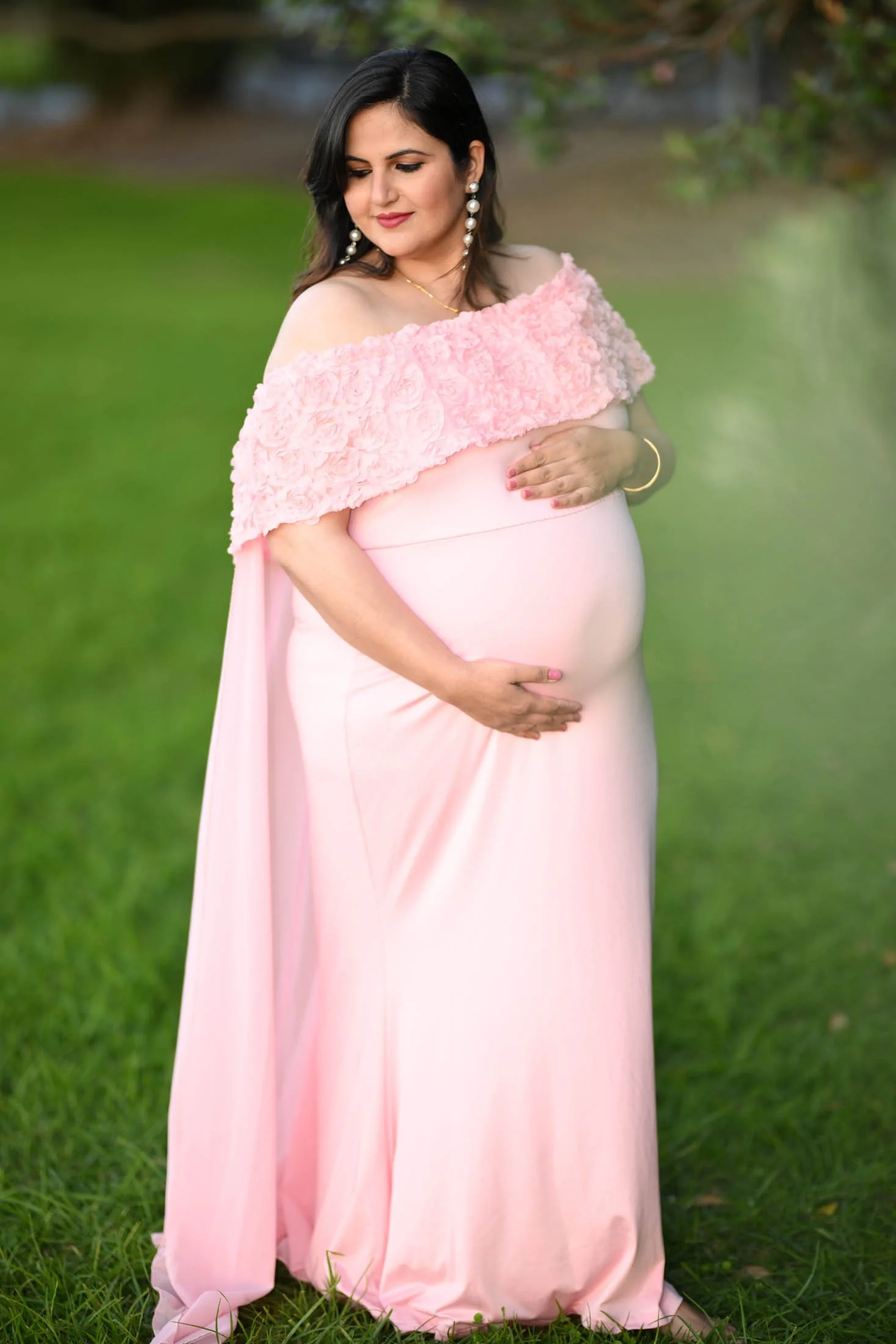 A pregnant woman in a pink gown with a floral shoulder cape, standing on green grass outdoors, gently holding her belly and looking down with a serene expression.