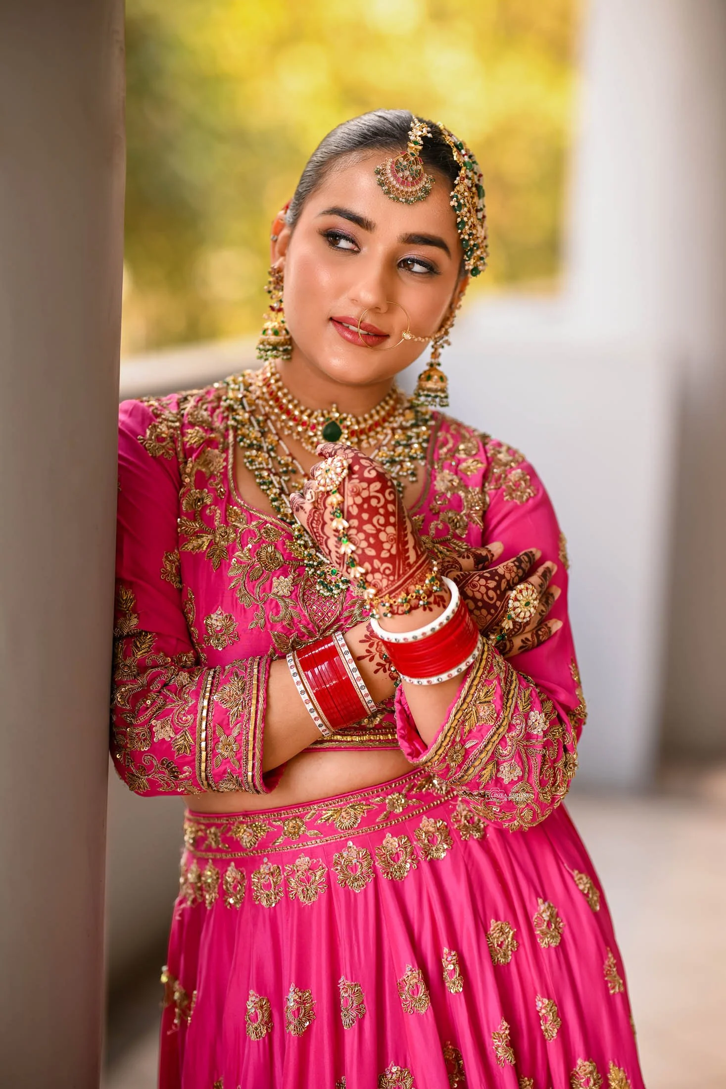 A woman dressed in traditional Indian bridal attire, wearing a bright pink outfit with gold embroidery, ornate jewelry including necklaces, earrings, and bangles, and henna designs on her hands, stands outdoors and looks thoughtfully to the side.
