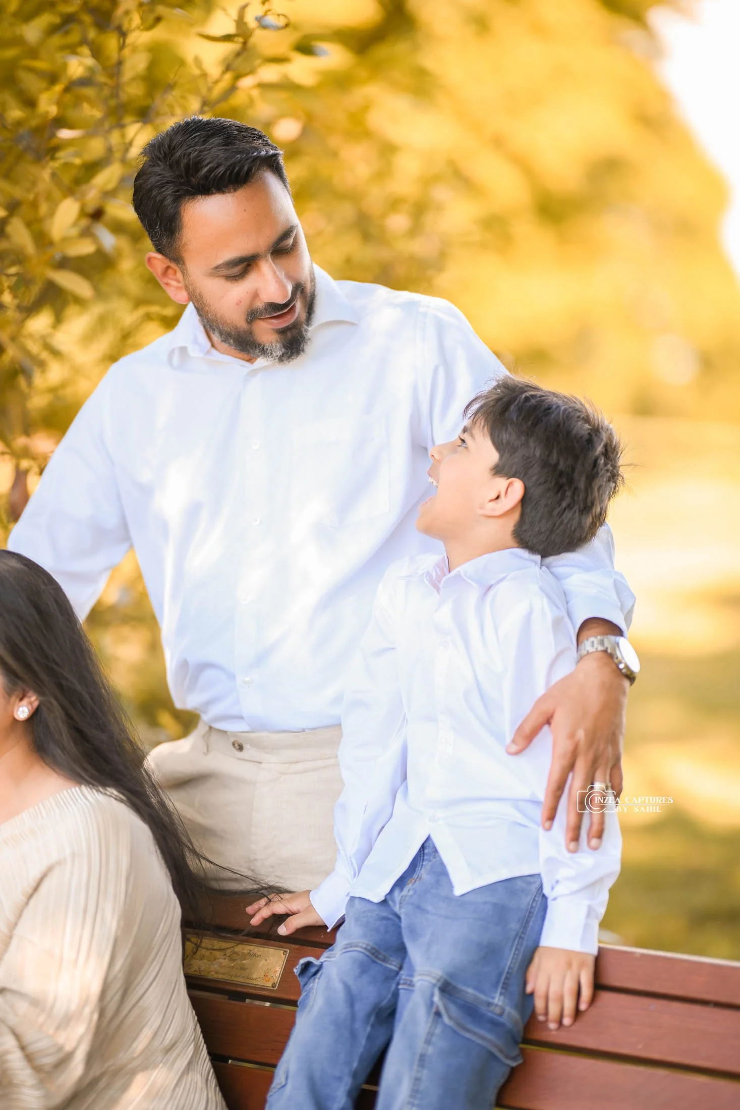 A man with a beard and a white shirt leaning on a park bench, smiling at a boy in a white shirt and jeans, who is also smiling and looking up at the man, with yellow autumn leaves in the background.