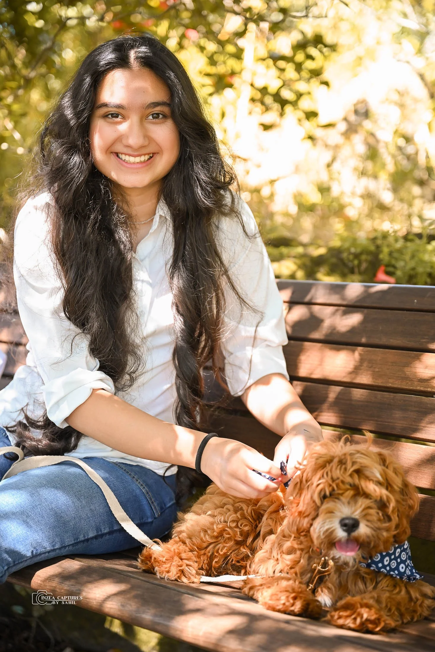 A young woman with long curly hair smiling while sitting on a wooden park bench. She is wearing a white shirt and blue jeans, and is holding a leash attached to a small, fluffy, reddish-brown dog with a blue bandana, sitting beside her with its tongu