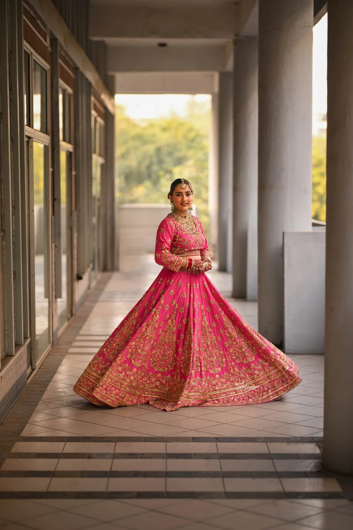 A woman dressed in a vibrant pink and gold traditional Indian bridal outfit, standing in an outdoor corridor with columns and glass doors.