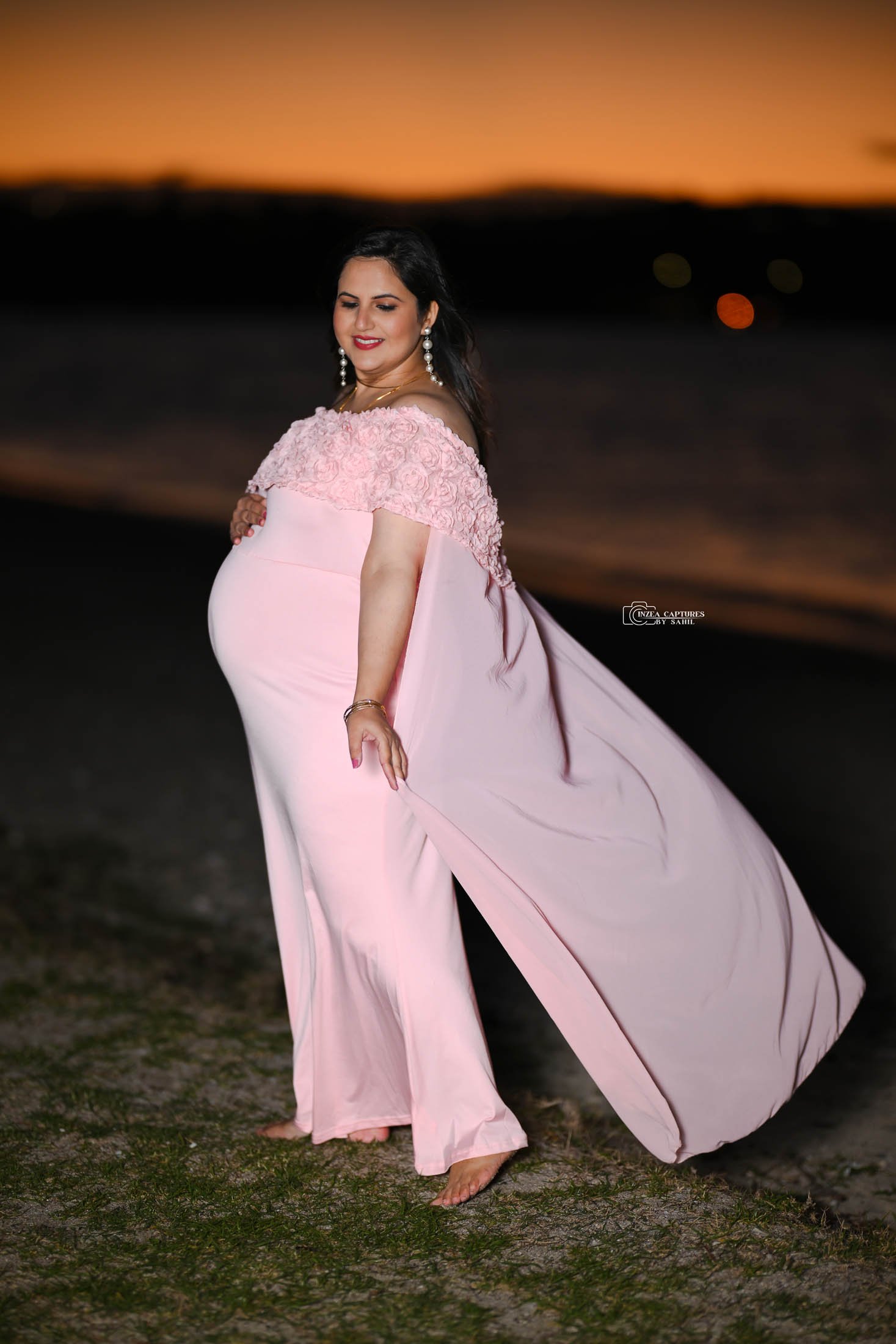 A pregnant woman in a pink flowing dress with floral details on the top, standing barefoot on a beach at sunset, smiling and looking down.