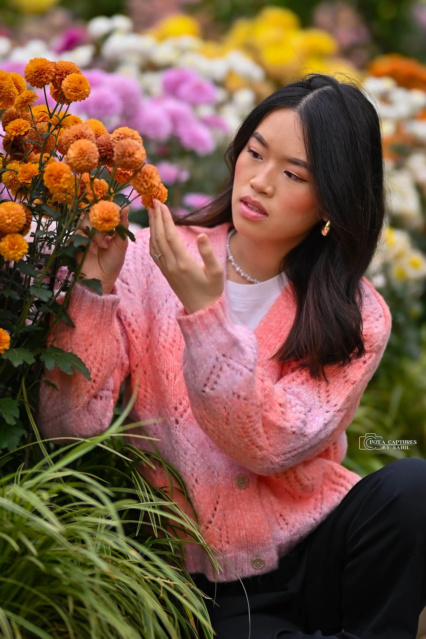 Young woman with dark hair, wearing a pink knit sweater, white top, and pearl necklace, gently touching orange marigold flowers in a colorful flower garden.