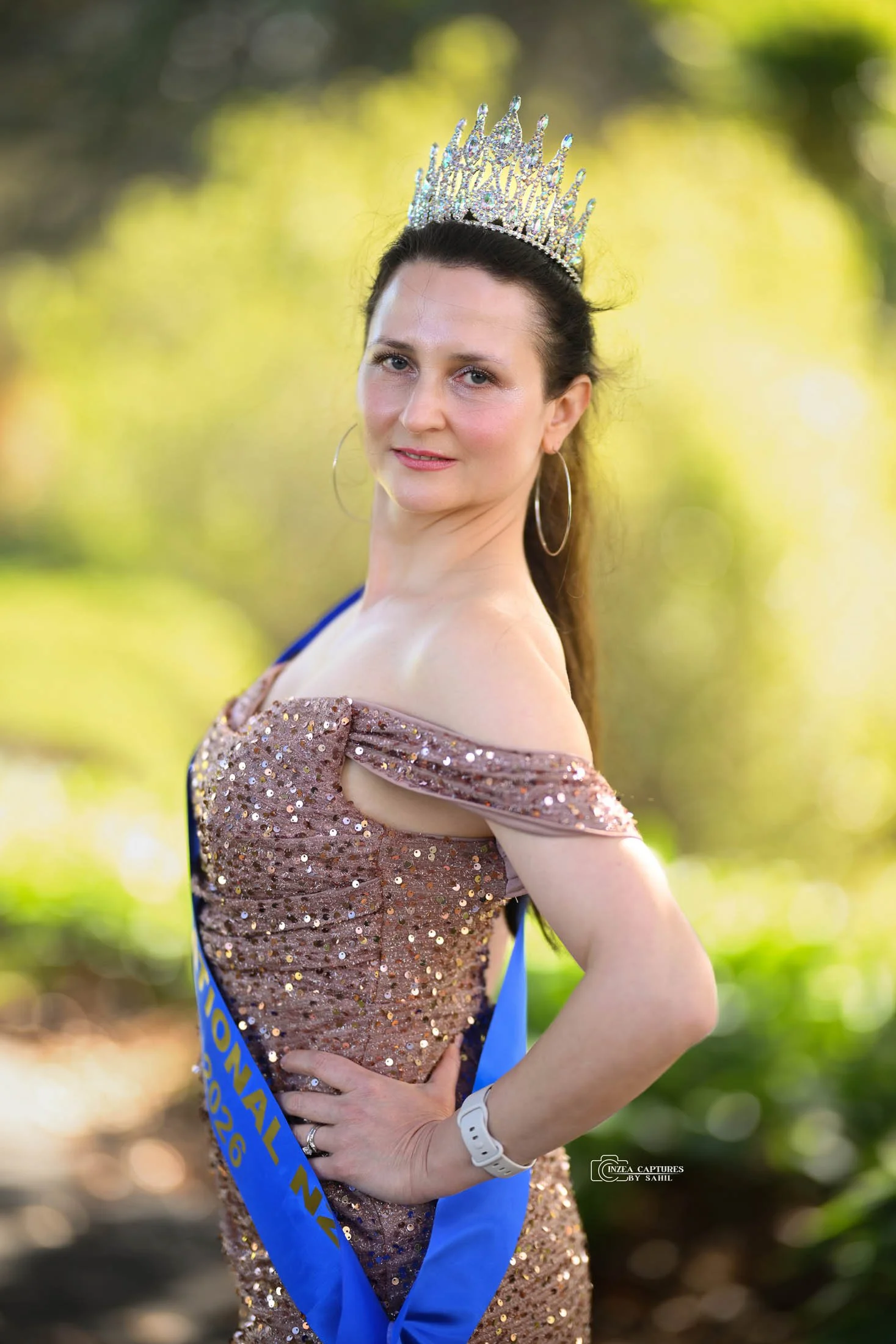 A woman wearing a crown and an off-the-shoulder, sequined dress, standing outdoors with a blurred green and yellow background.