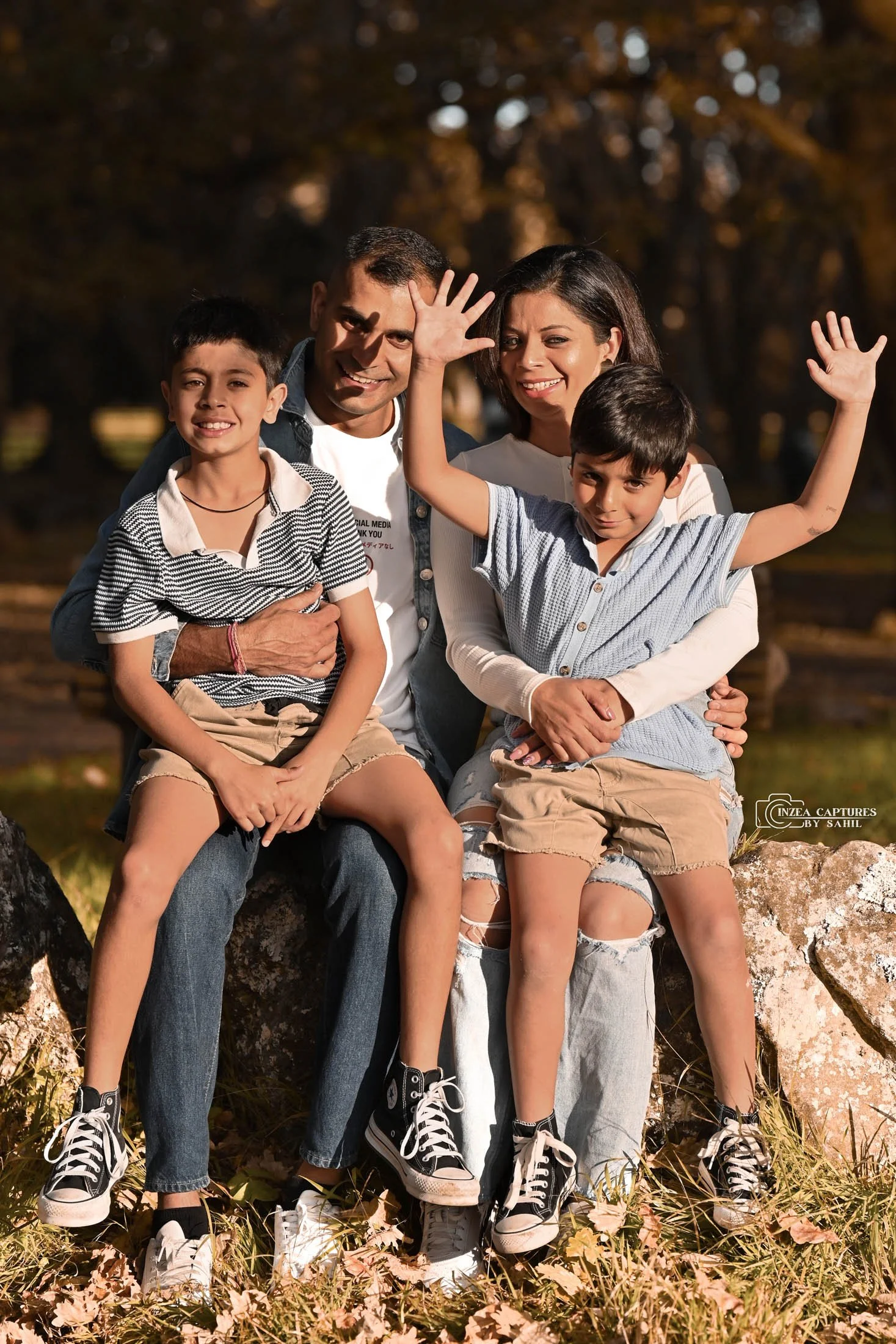 Family sitting on rocks outdoors, smiling and waving. Three children and two adults, fall foliage in background, casual clothing, some wear sneakers.