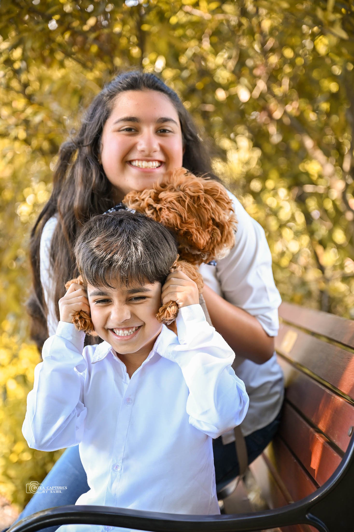 A young girl and boy sitting on a bench outdoors, smiling, with a fluffy brown dog between them. The girl is hugging the dog, and the boy is holding dog ears on his head, pretending to be a puppy.