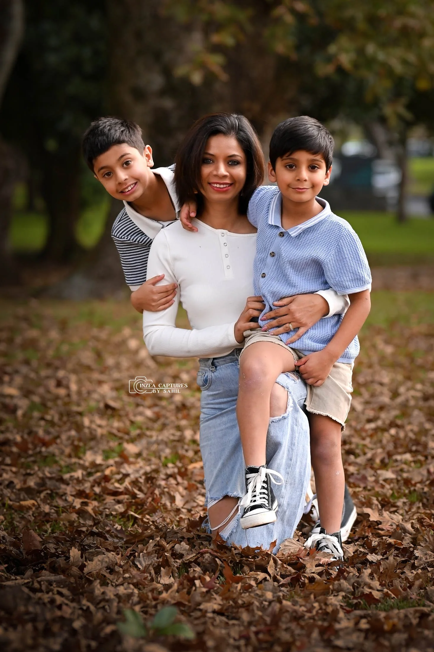 A woman and two young boys outdoors in a park during fall, smiling at the camera. The woman is kneeling on the ground with one boy sitting on her knee and the other boy standing behind her, with their arms around her.