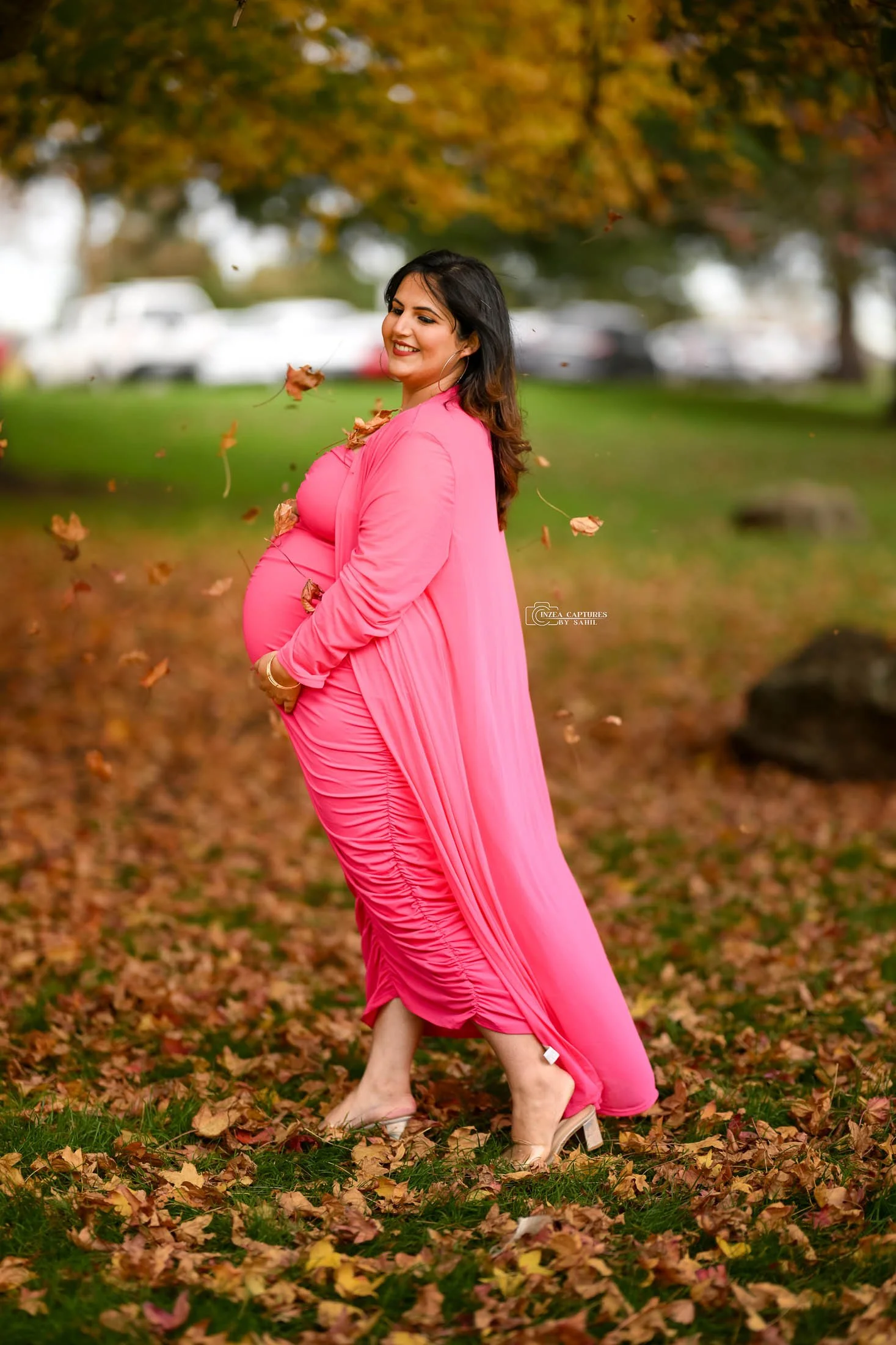 Pregnant woman in a pink dress standing on a leaf-covered ground in a park, smiling and holding her belly, with falling leaves around her and colorful autumn trees in the background.