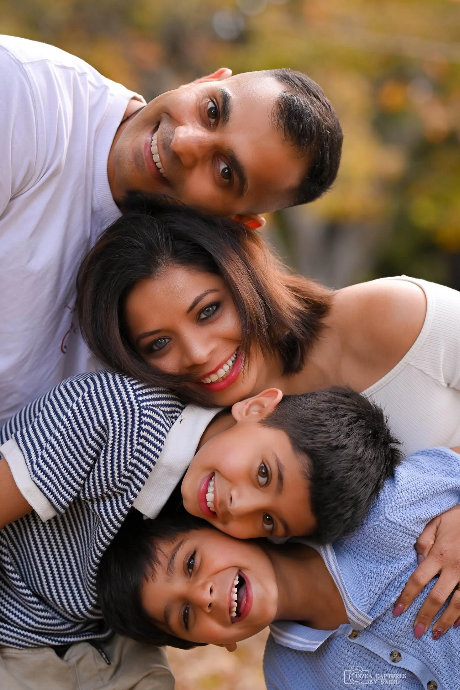 A smiling family of four lying outdoors in autumn, with a man, a woman, and two boys, all hugging and looking at the camera.