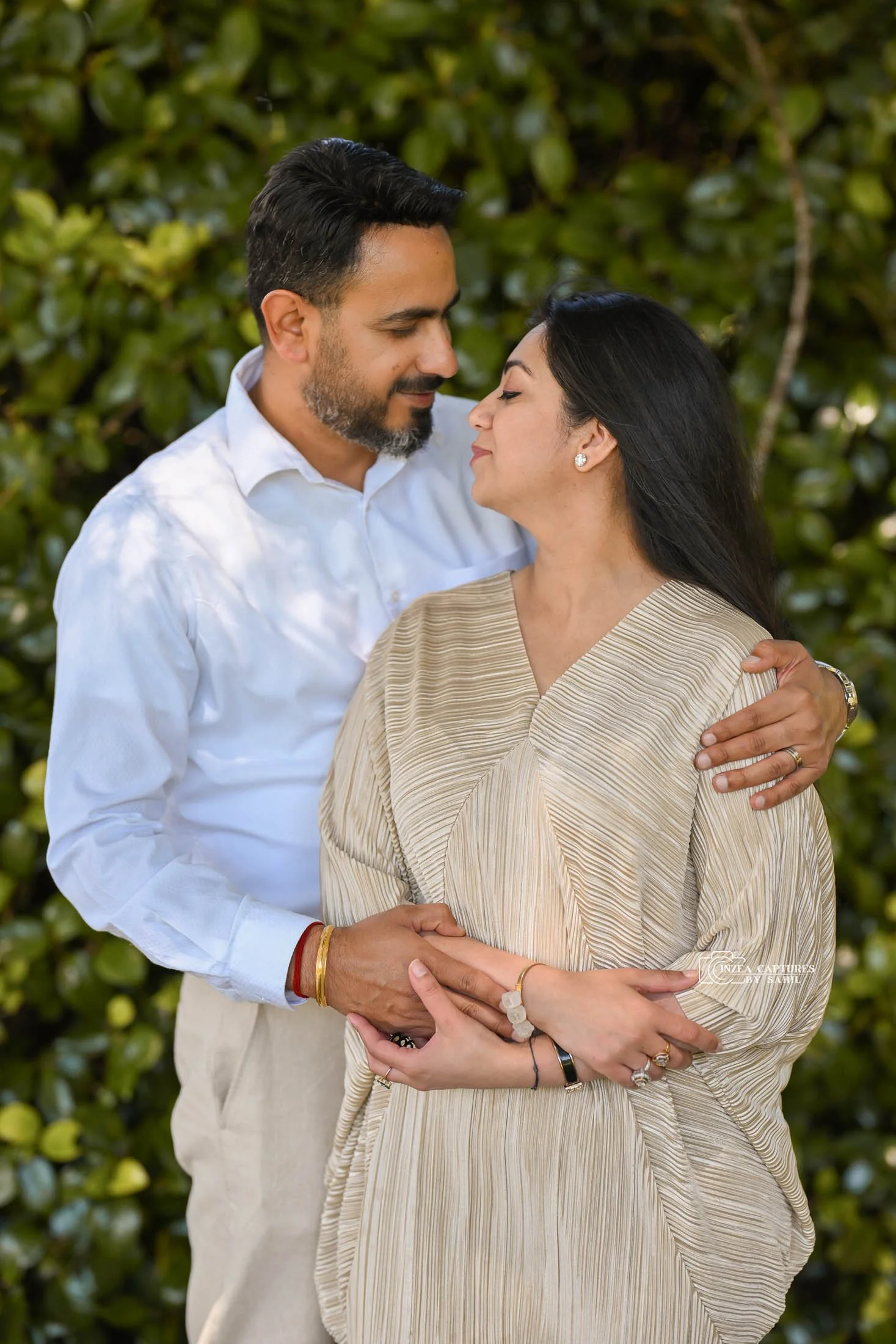 A couple standing close together, looking at each other affectionately, outdoors with green foliage in the background.