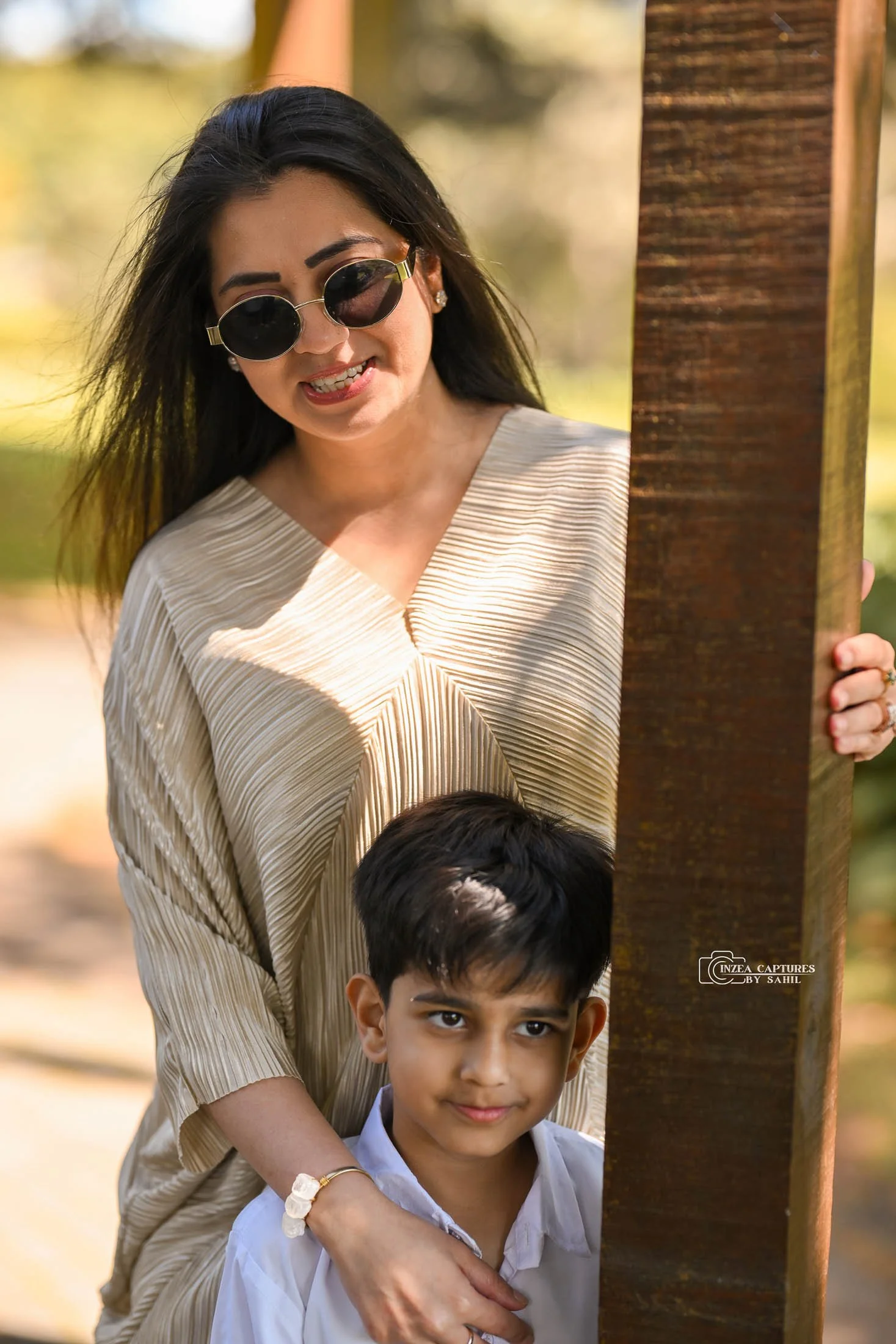 A woman with long black hair and sunglasses smiling, standing behind a young boy with short black hair, both peeking out from behind a vertical wooden post outdoors on a sunny day.