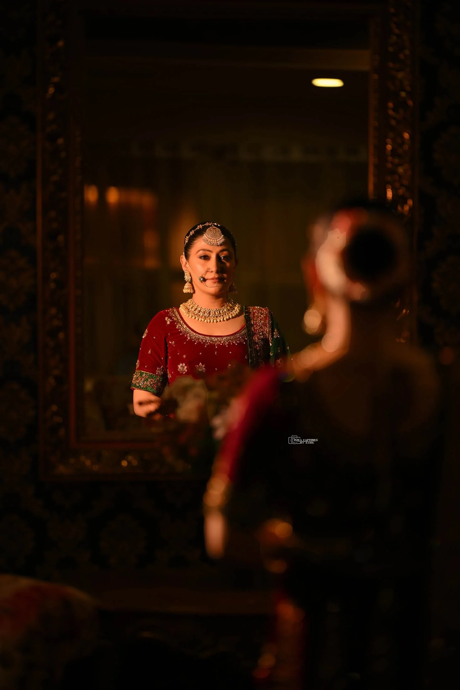 A woman dressed in traditional Indian bridal attire is looking at herself in a mirror. She is wearing ornate jewelry including earrings, a necklace, and a headpiece, with a red and green embroidered outfit.