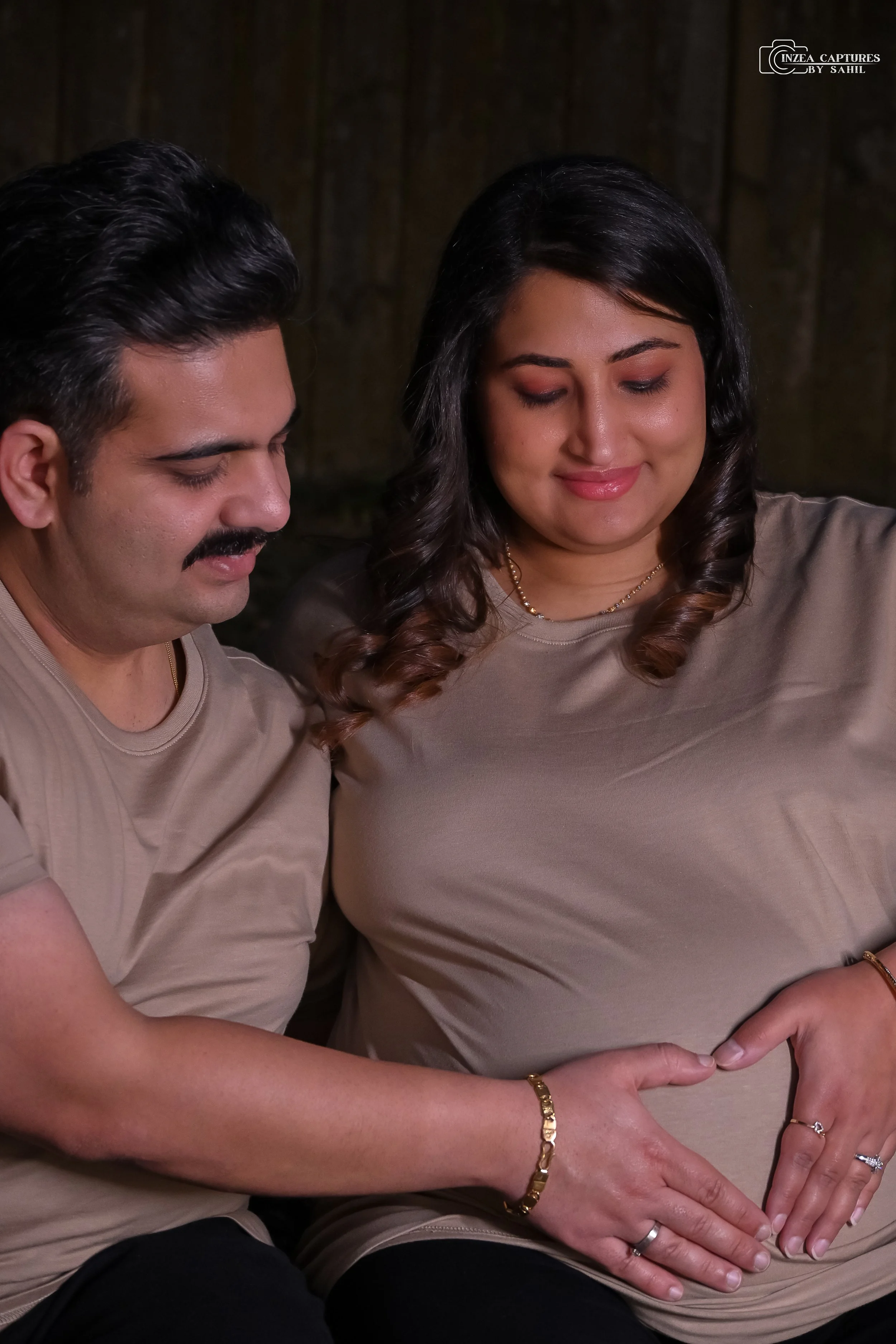 A man and a pregnant woman sit closely together, smiling softly and touching her pregnant belly with their hands.