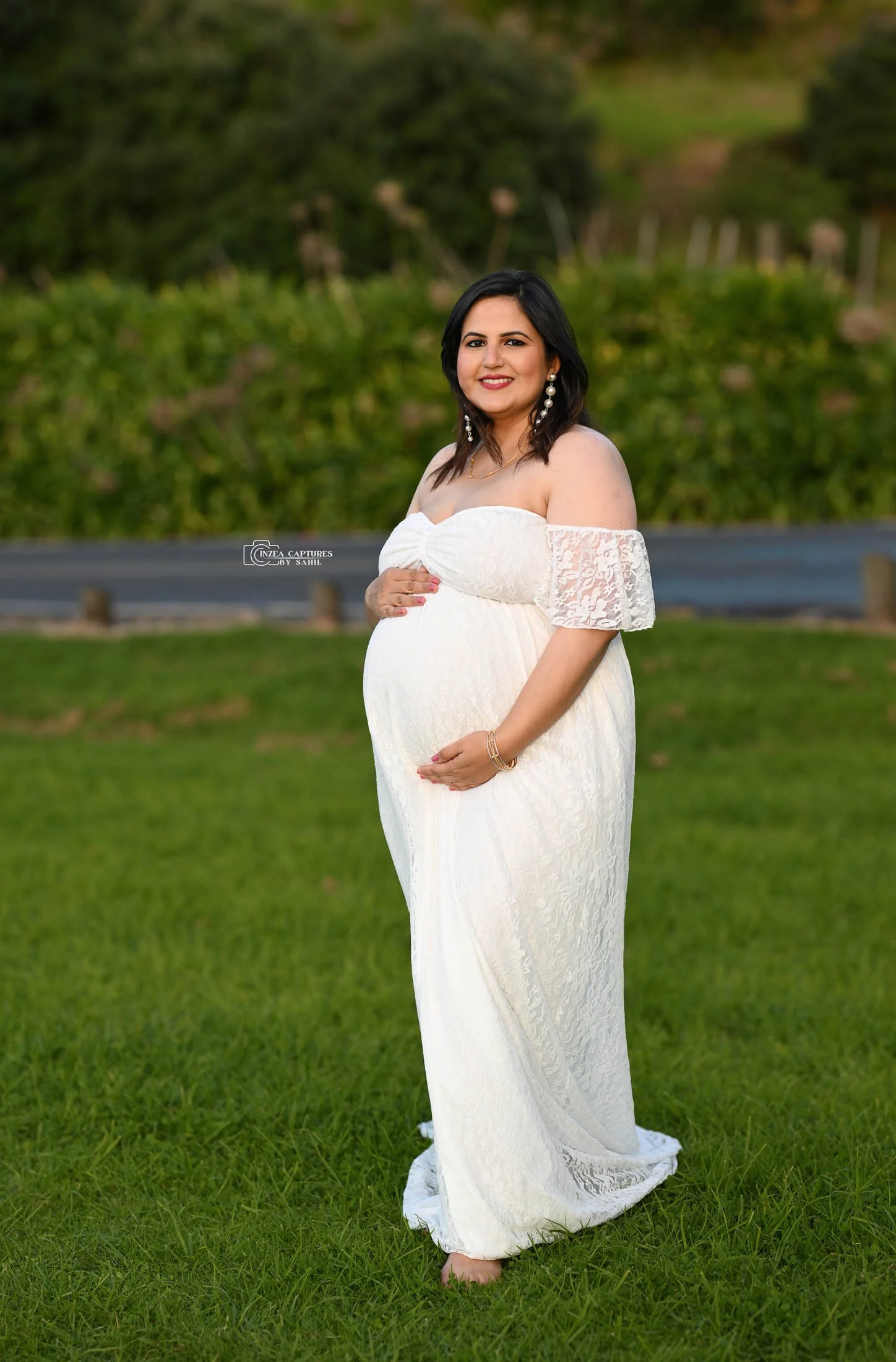 A pregnant woman in a white lace dress stands on grass with green trees in the background, smiling and holding her belly.