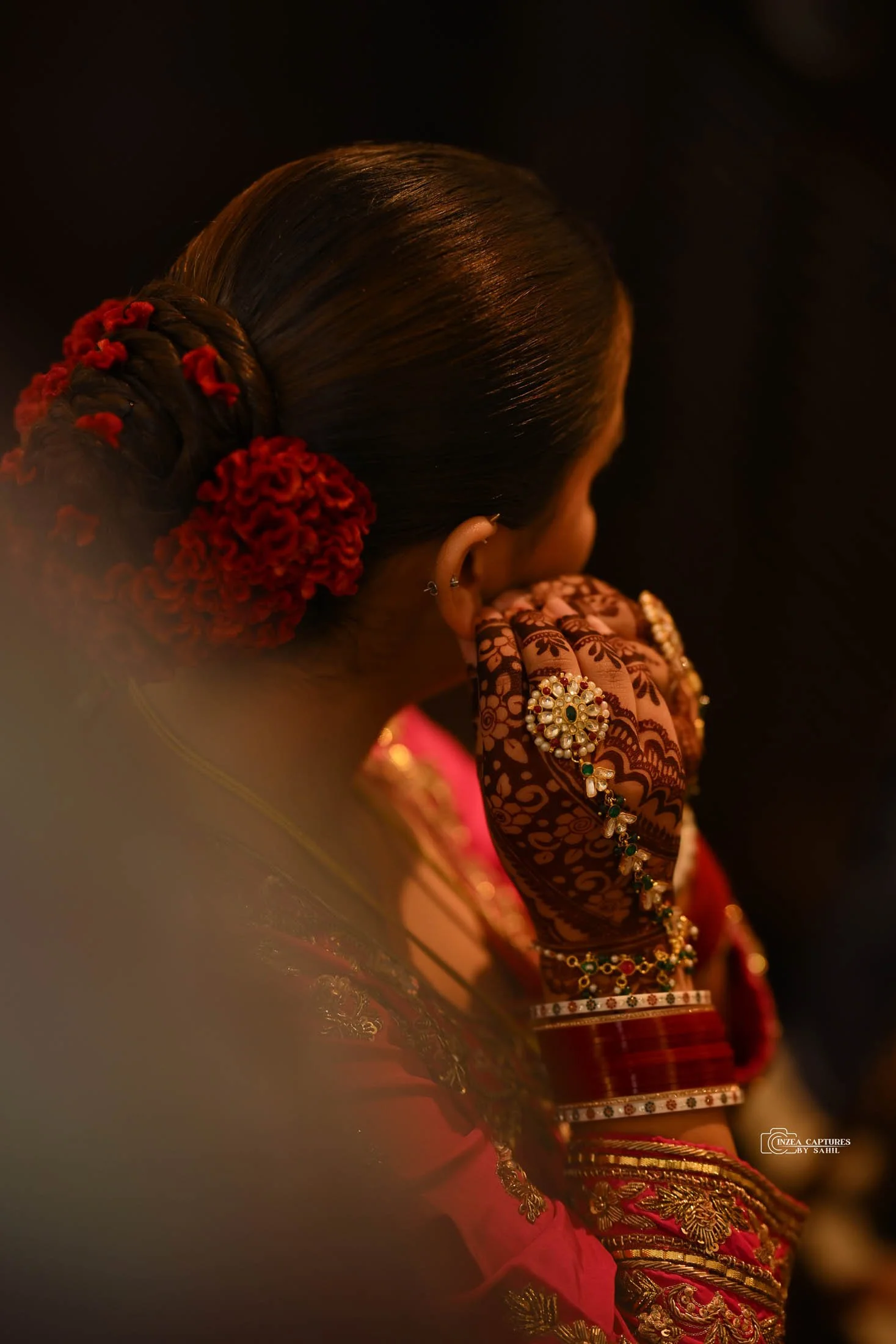 A woman with elaborately styled hair adorned with red flowers, wearing traditional Indian jewelry and clothing, covering her face with her hand decorated with henna and jewelry.